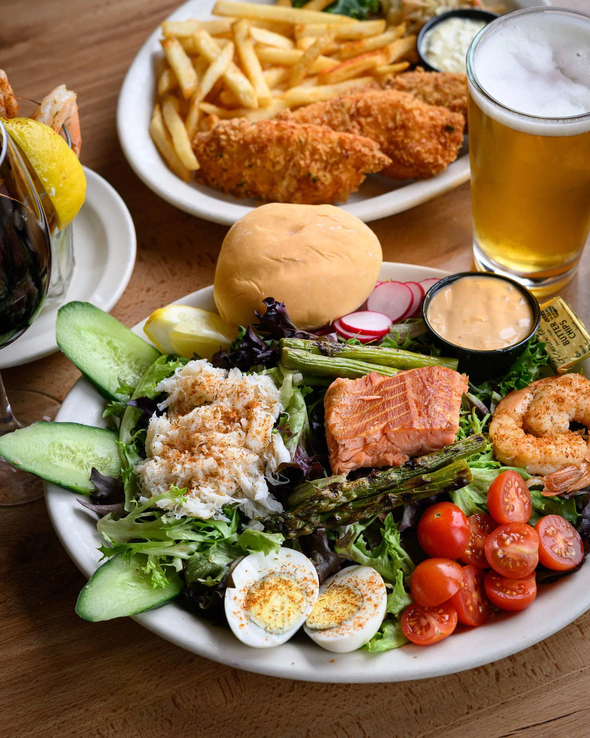 A plate of fresh salad with cherry tomatoes, boiled eggs, cucumbers, and greens, accompanied by grilled salmon, asparagus, and a bun, served on a wooden table.