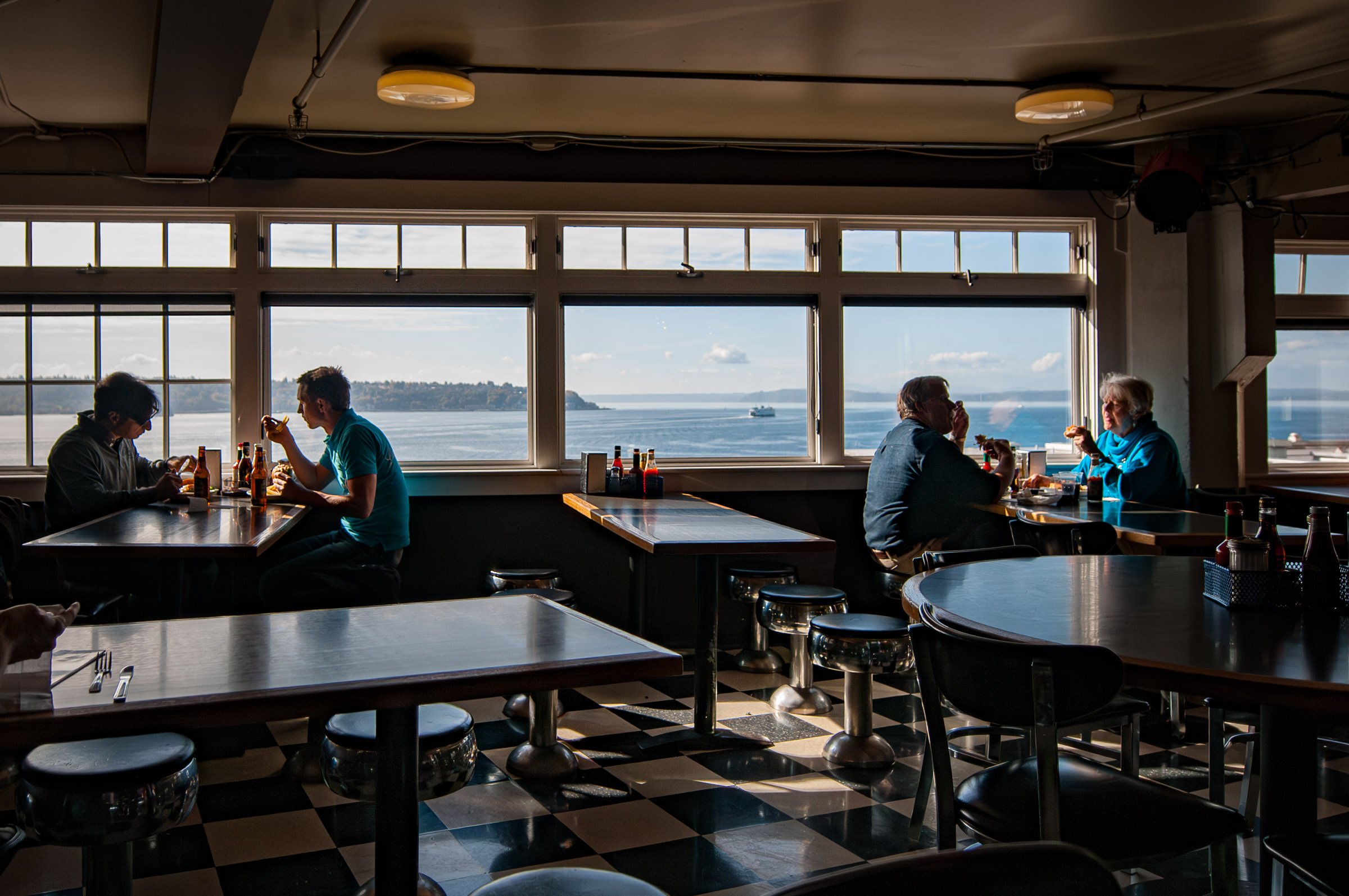 A restaurant interior with large windows overlooking a body of water and boats, with four people sitting at tables, eating and talking.