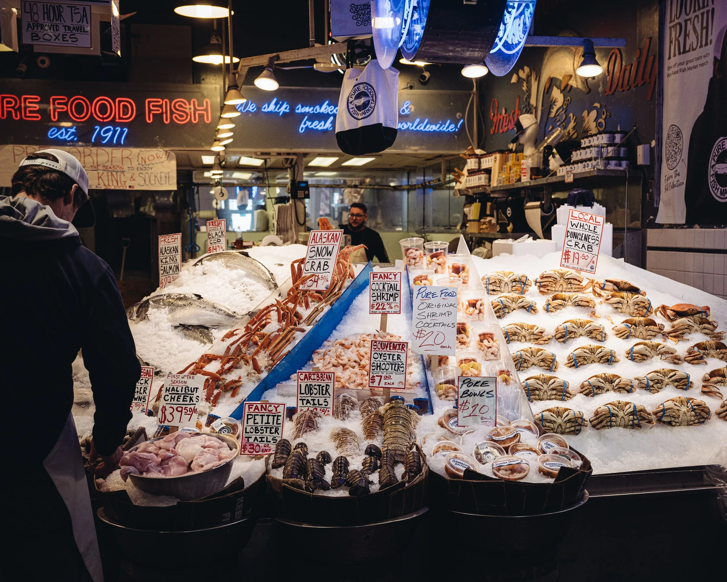 Display of fresh seafood at a market stall, including crabs, lobster tails, shrimp, and fish, with prices and labels. Overhead neon lights illuminate the scene with signs indicating the market's history and offerings.