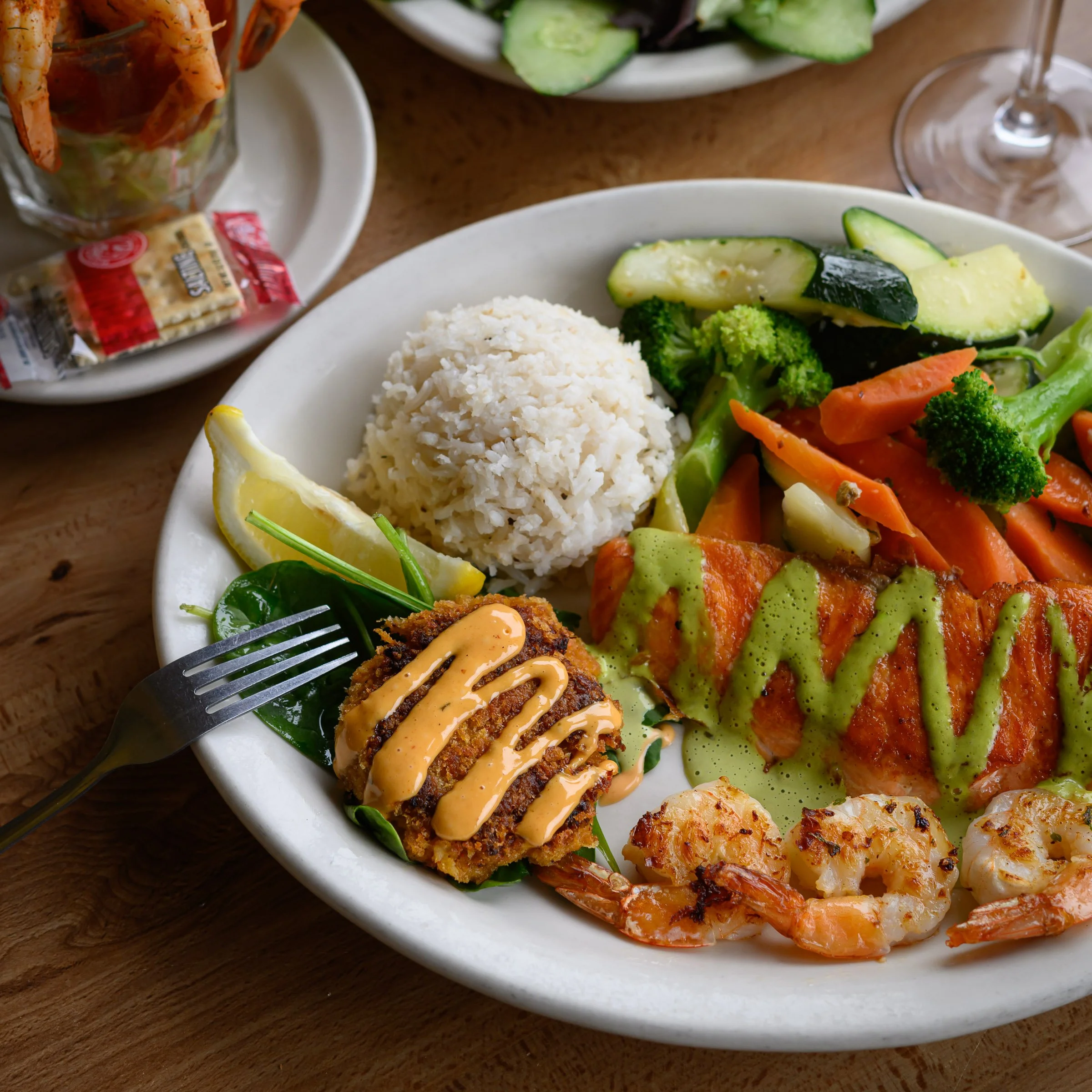 Close-up of a white oval plate with cooked rice, grilled shrimp, vegetables, and a lemon wedge. In the background, a glass of white wine, a small bowl of salad, and a small dish of baked appetizer with orange sauce are visible on a wooden table.