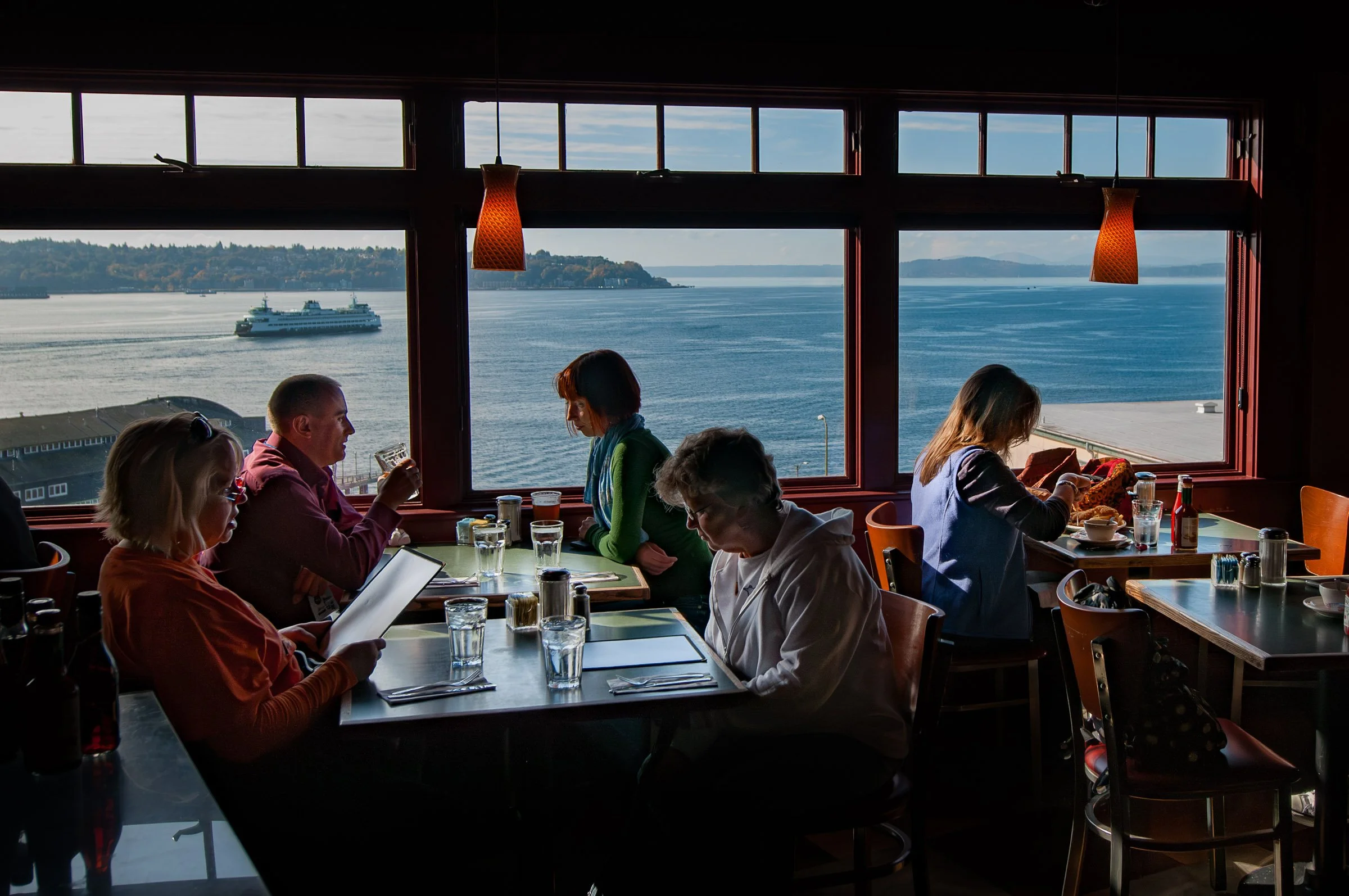 People dining at a restaurant with large windows overlooking the water and a ferry boat passing by during daytime.