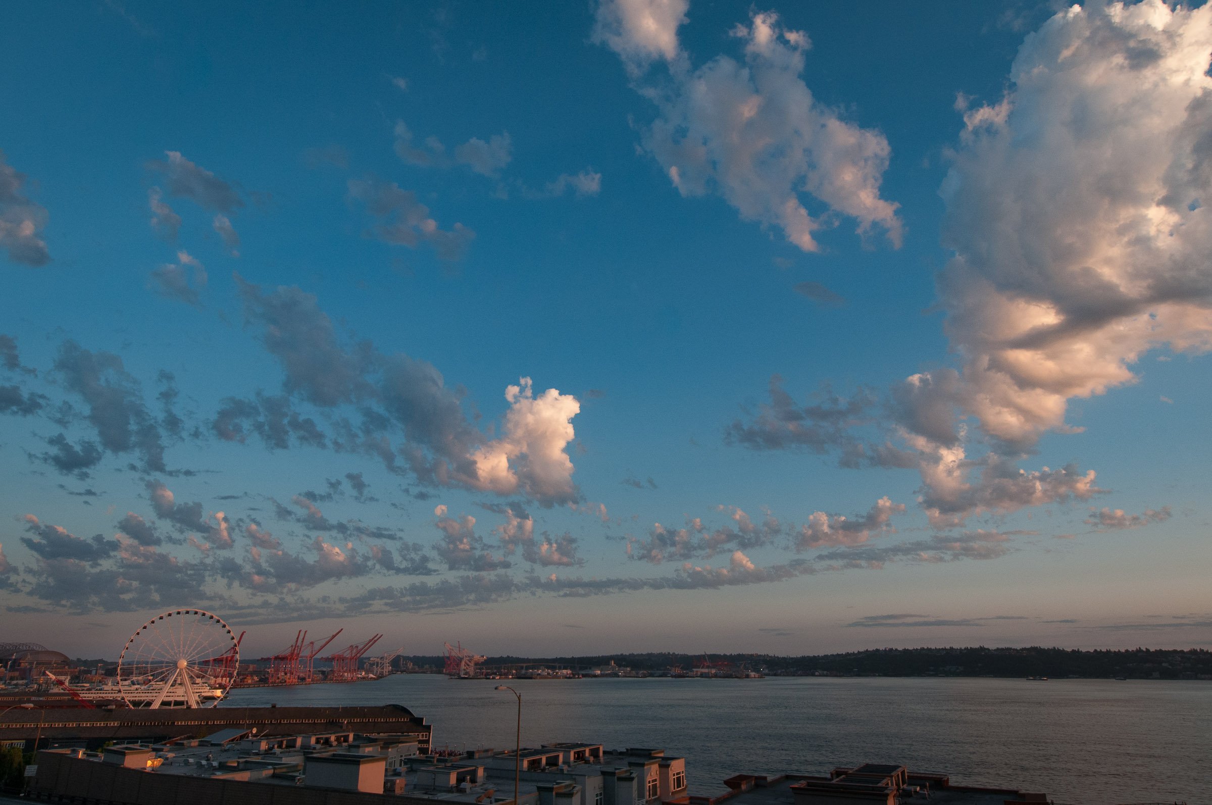 A panoramic view of a sky filled with scattered clouds during sunset above a city waterfront, with a Ferris wheel and cranes in the background.