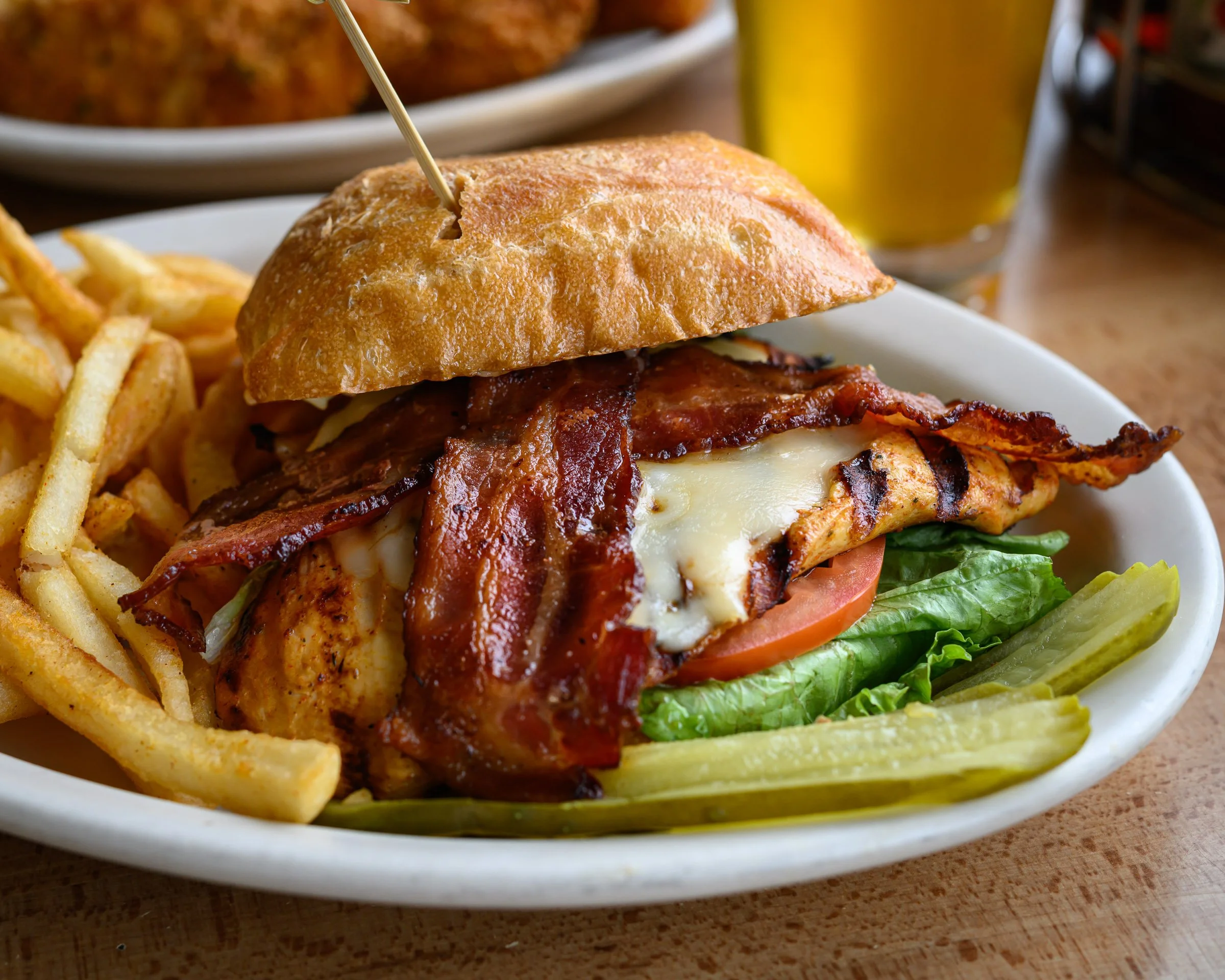 Close-up of a cheeseburger with bacon, lettuce, tomato, and cheese in a toasted bun, served on a white plate with French fries and a glass of yellow beverage in the background.