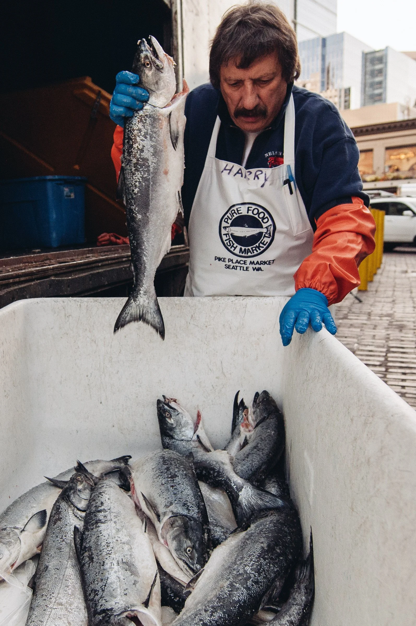 A man with a mustache, wearing a white apron with the words 'Pure Food Fish Market' and gloves, holding a large fish at a seafood market with other fish in a white container and tall buildings in the background.