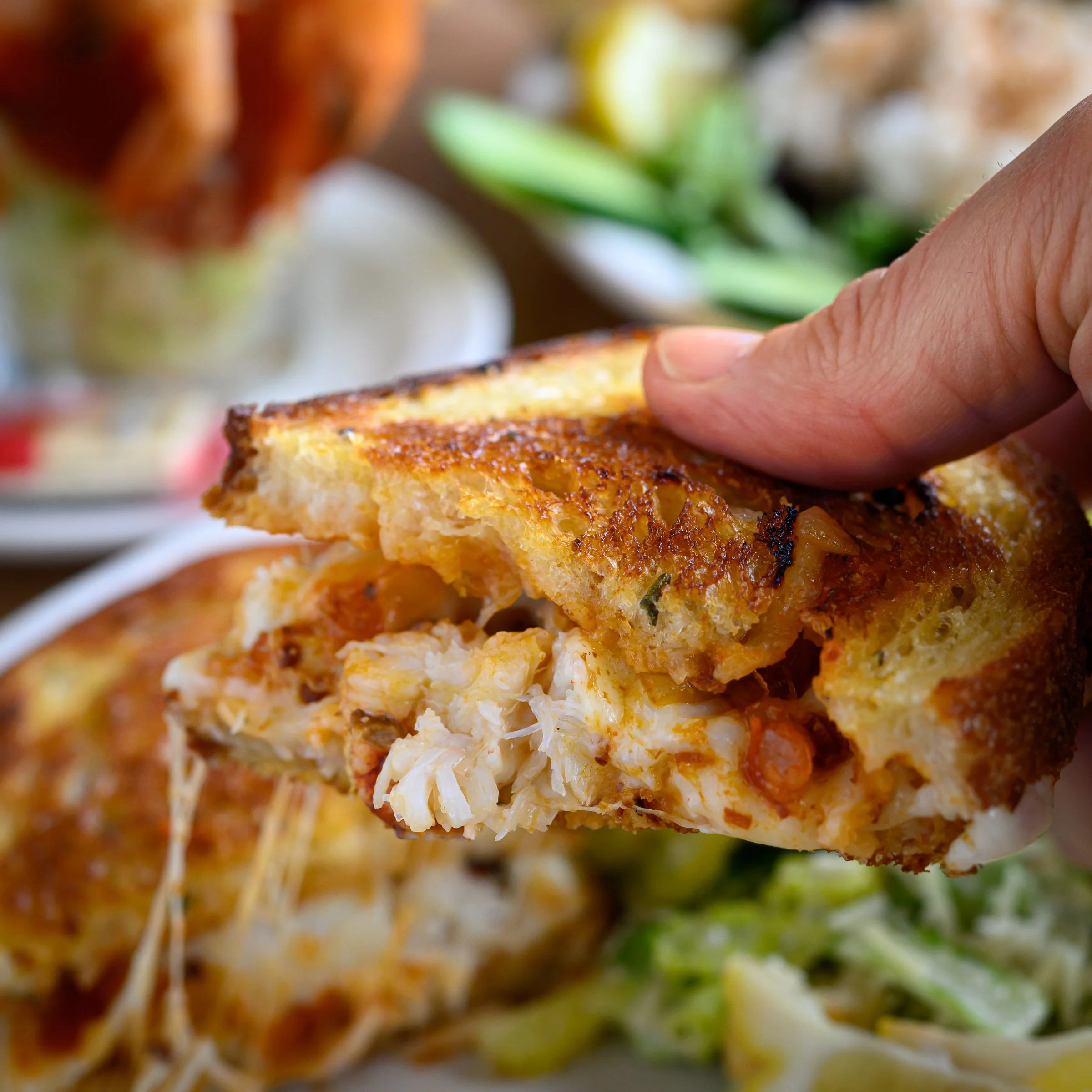 Close-up of a cooked crab cake held by a person's hand, with a blurred background of other crab cakes and green garnish.