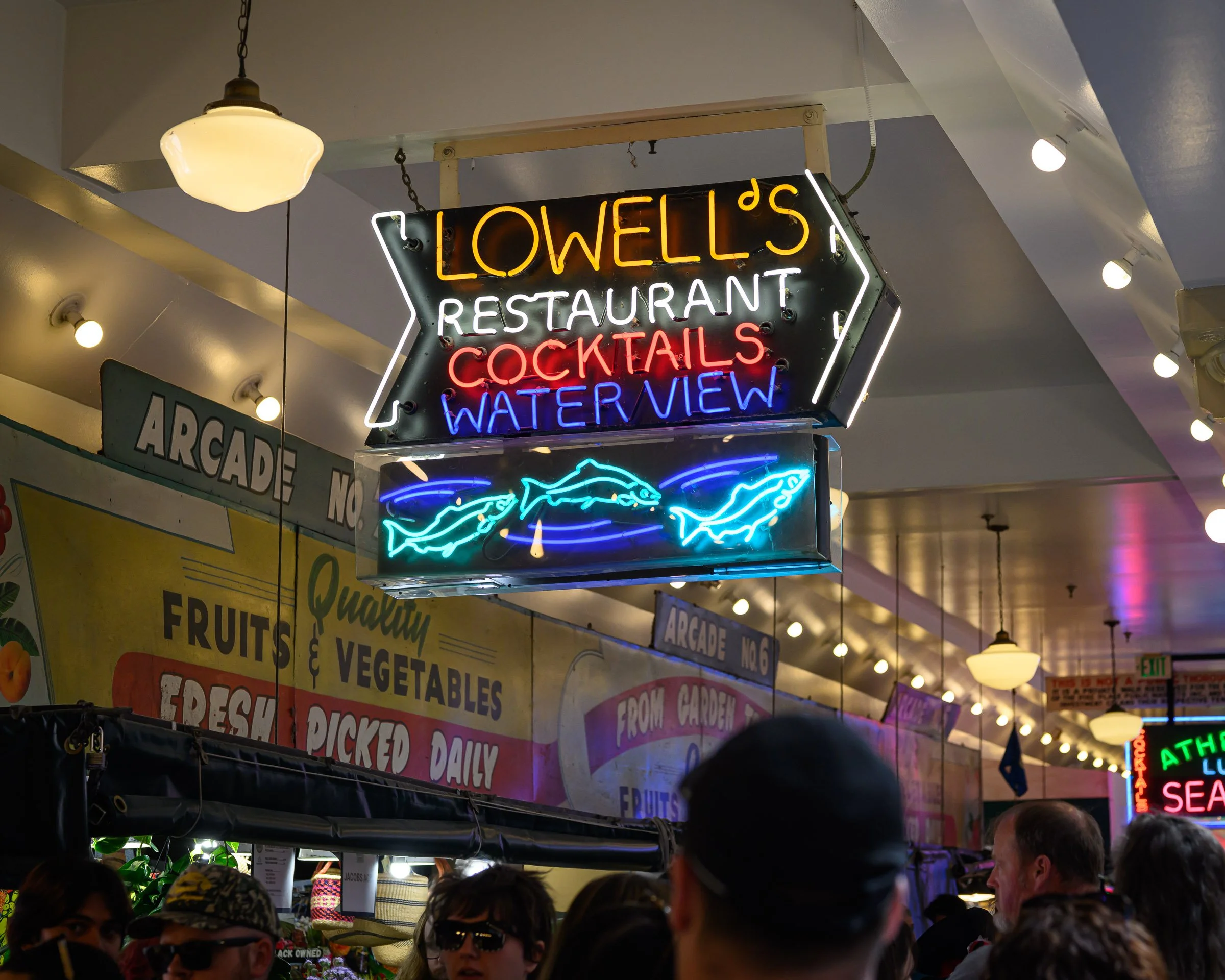 Neon sign reading 'Lowell's Restaurant Cocktails Waterview' hanging from ceiling at busy indoor market with people and display booths.