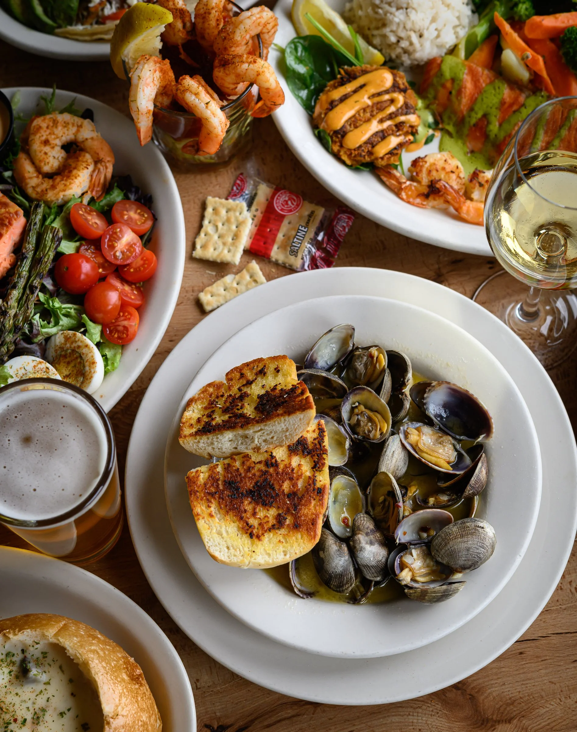 A variety of food dishes on wooden table including clam pasta, grilled toast with tomato, salad with cherry tomatoes, shrimp cocktail, fried fish, and a glass of white wine.