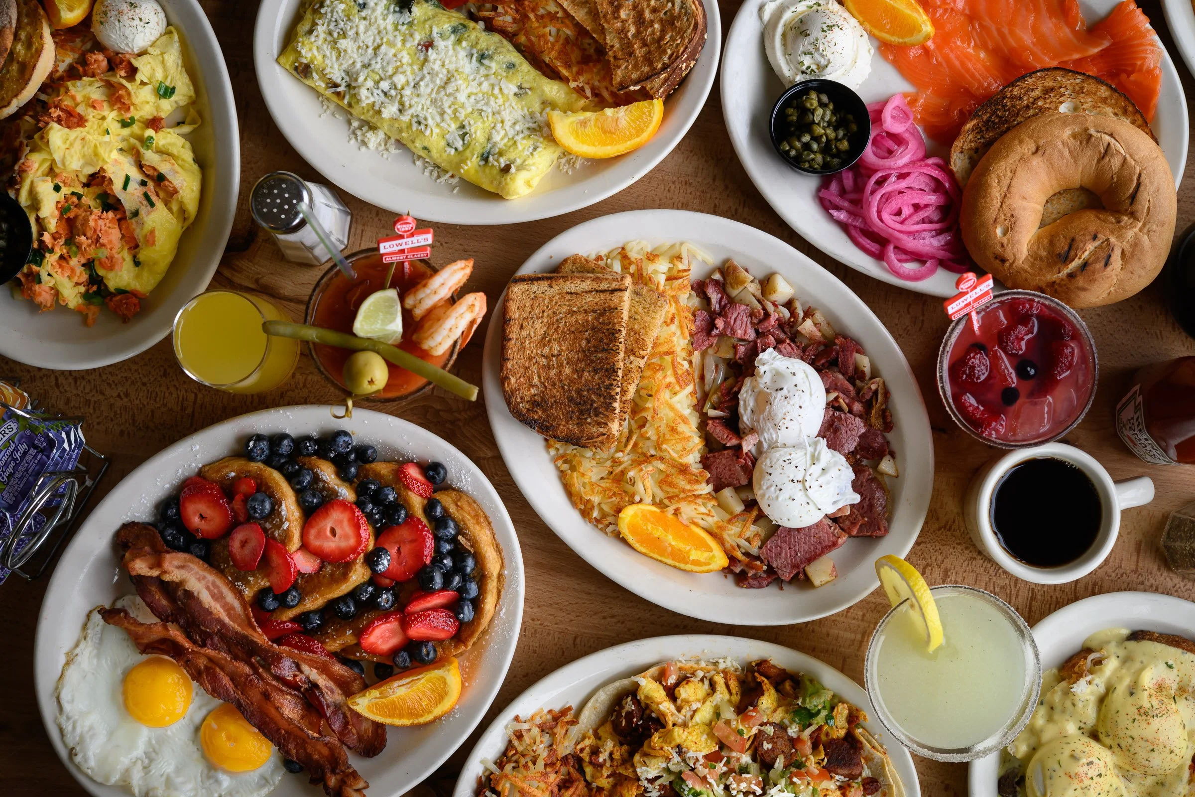A variety of breakfast dishes including fried eggs with bacon, fruit-topped pancakes, scrambled eggs, smoked salmon with bagels, breakfast pizza, and assorted sides like fried potatoes, pickles, and pink onions.
