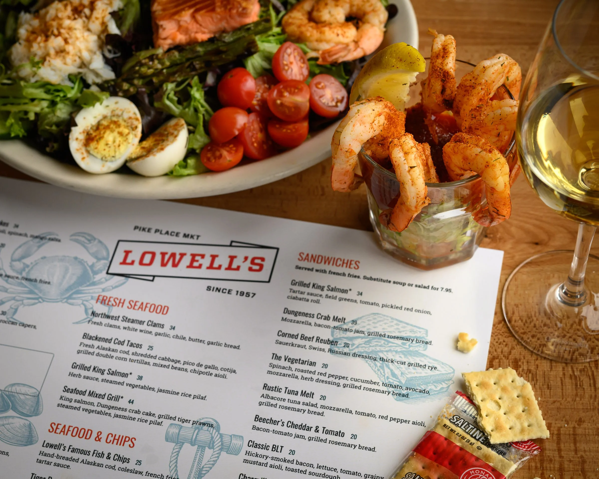 A restaurant menu from Lowell's listing seafood, chicken, and other dishes, partially visible under a plate of salad with cherry tomatoes, boiled eggs, and grilled salmon, alongside a glass of white wine and a few crackers.