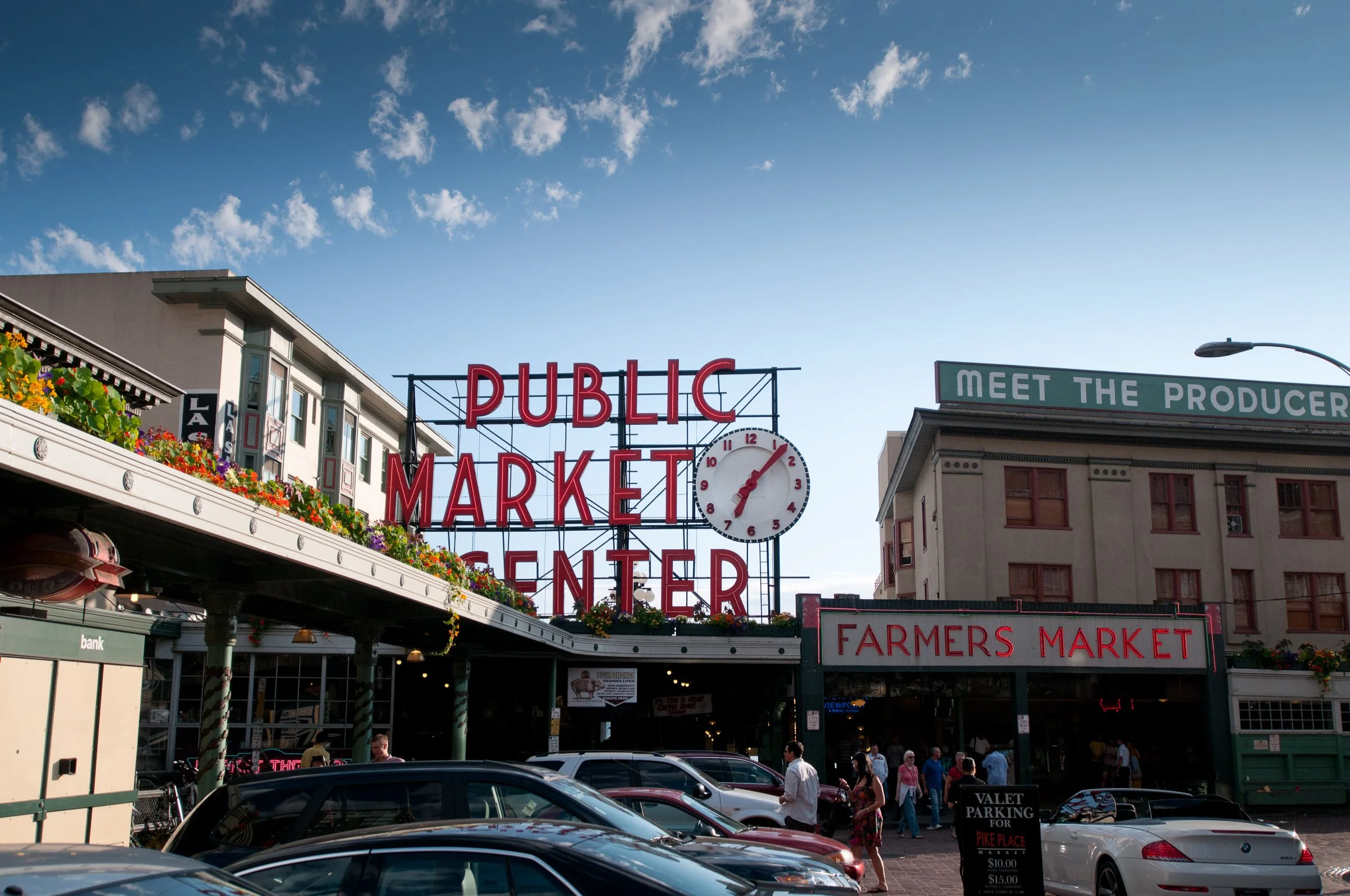 View of Pike Place Market in Seattle with a large red sign that reads 'Public Market Center' and a clock, buildings, pedestrians, and cars in the foreground, under a partly cloudy sky.