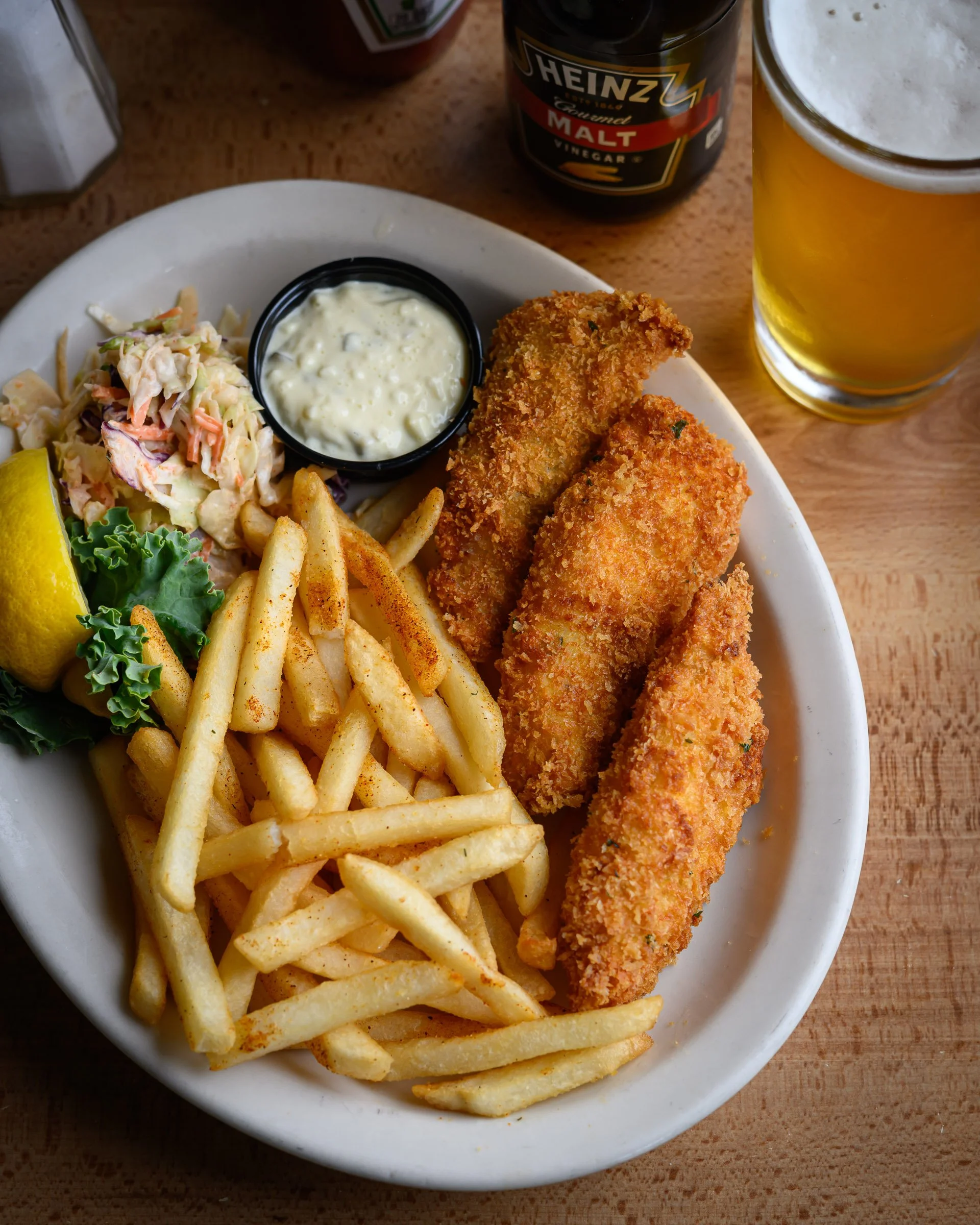 A plate with French fries, coleslaw, and two pieces of fried fish fillets served with tartar sauce, alongside a lemon wedge, with a glass of beer and a bottle of Heinz malt vinegar in the background.