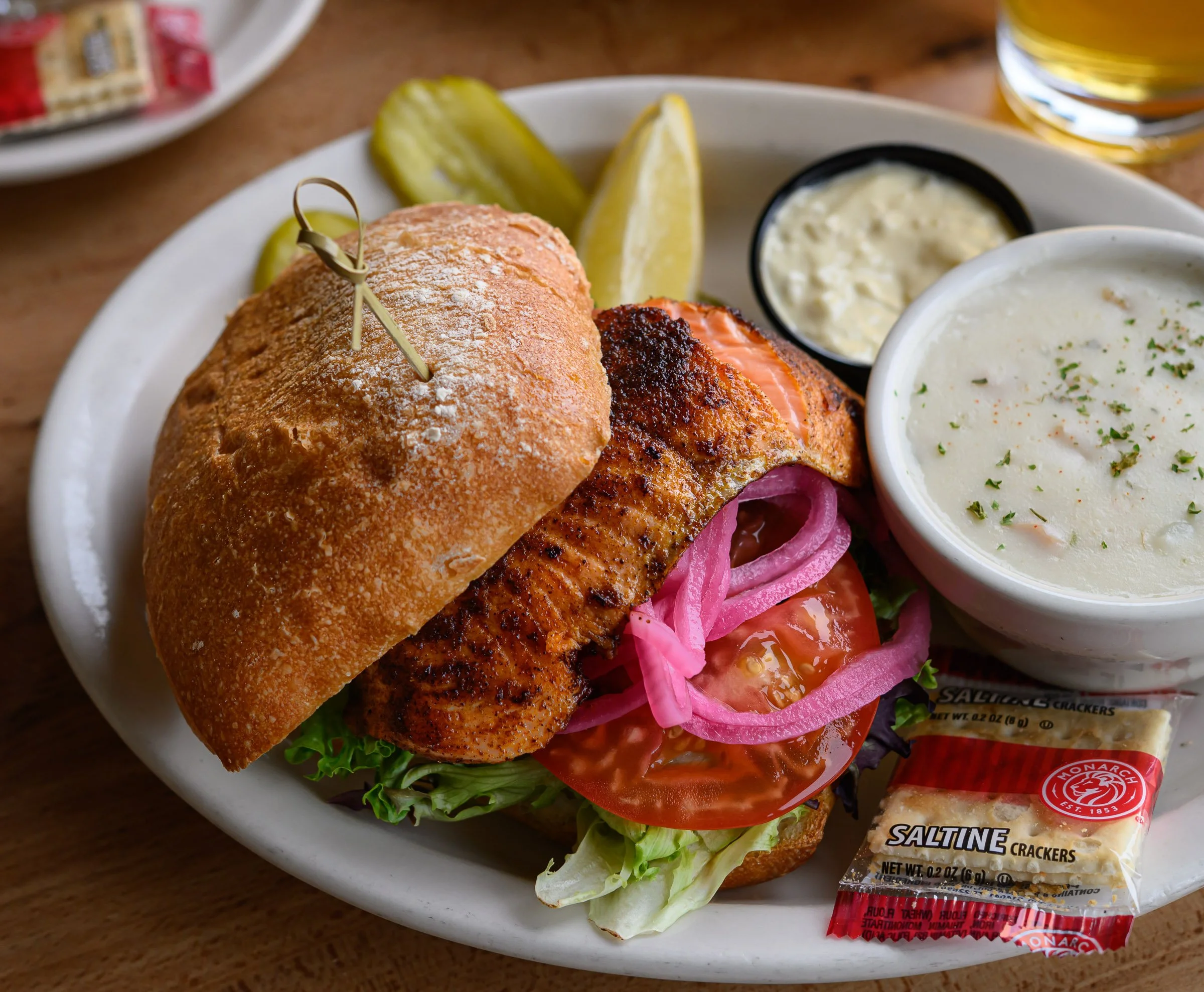 A plate of food with a sandwich containing grilled salmon, lettuce, tomato, and pickles, accompanied by two small bowls of sauces, surrounded by pickles and packets of saltine crackers.