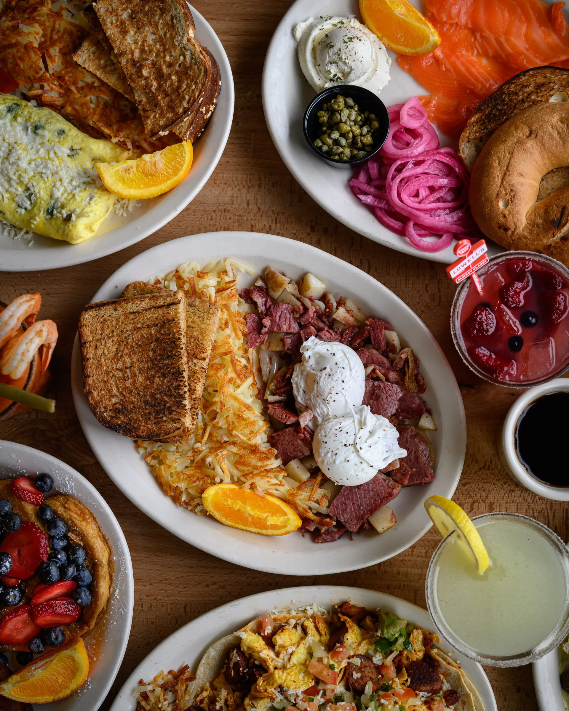 A variety of breakfast dishes including pancakes with berries, scrambled eggs with a slice of orange, bagels with smoked salmon, and a bowl of green syrup, all on white plates on a wooden table.