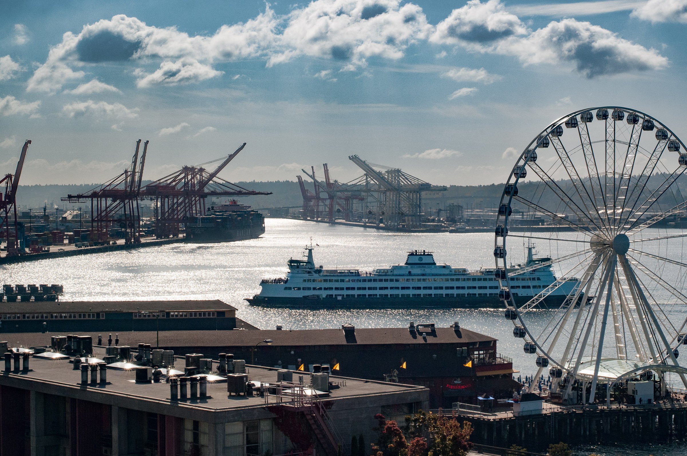 A harbor scene with a large Ferris wheel on the right, cruise ships, and numerous cargo cranes along the waterfront with a partly cloudy sky overhead.
