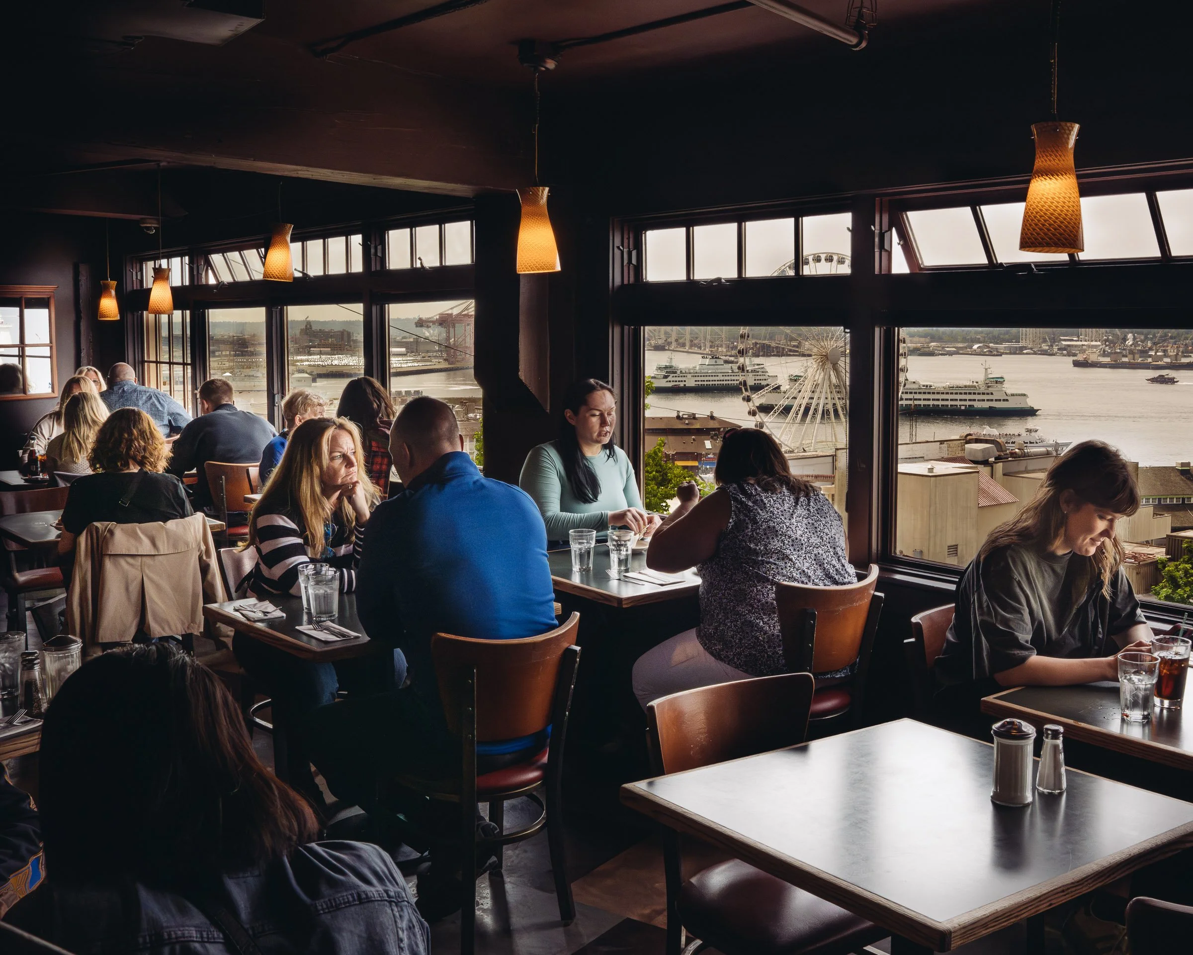 Indoor restaurant dining area with large windows overlooking a harbor with ships and a Ferris wheel. Several people are seated at tables, enjoying their meals with glasses of water and drinks. Warm hanging lights illuminate the space.