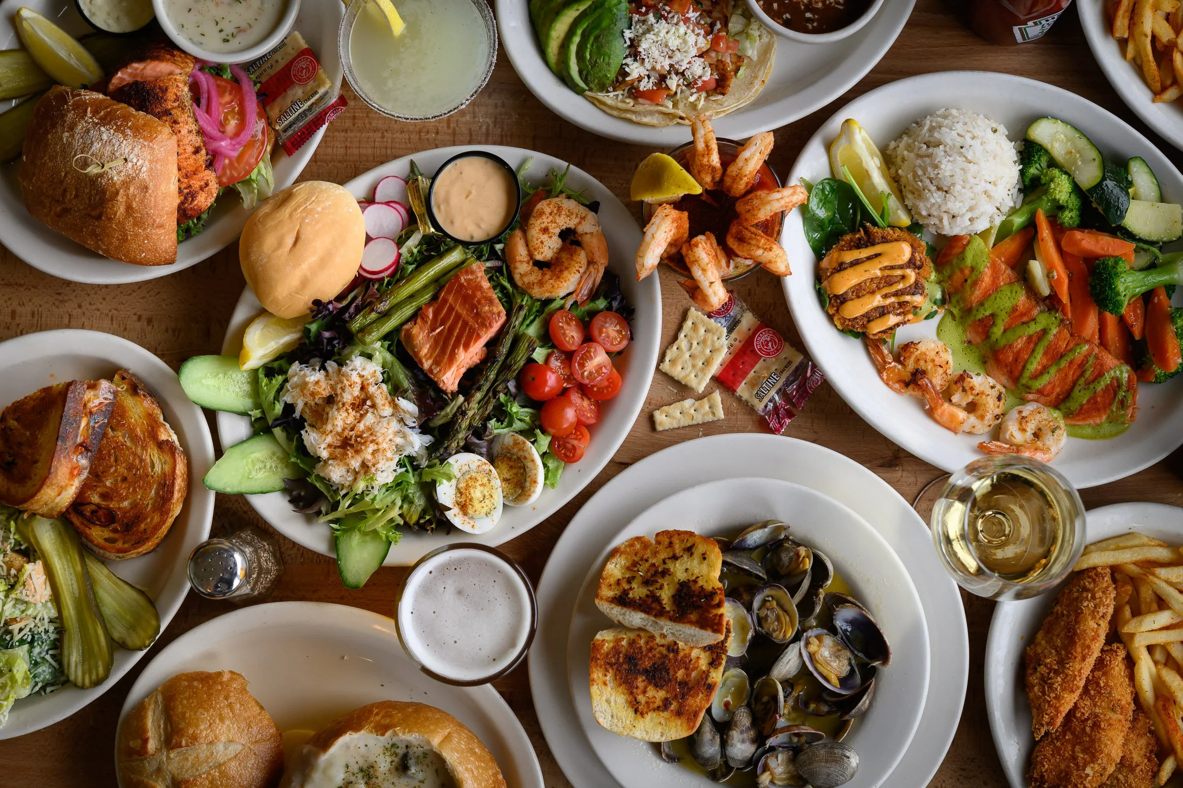 A variety of plates filled with food, including salads, seafood, and fried dishes, along with drinks and condiments on a wooden table.