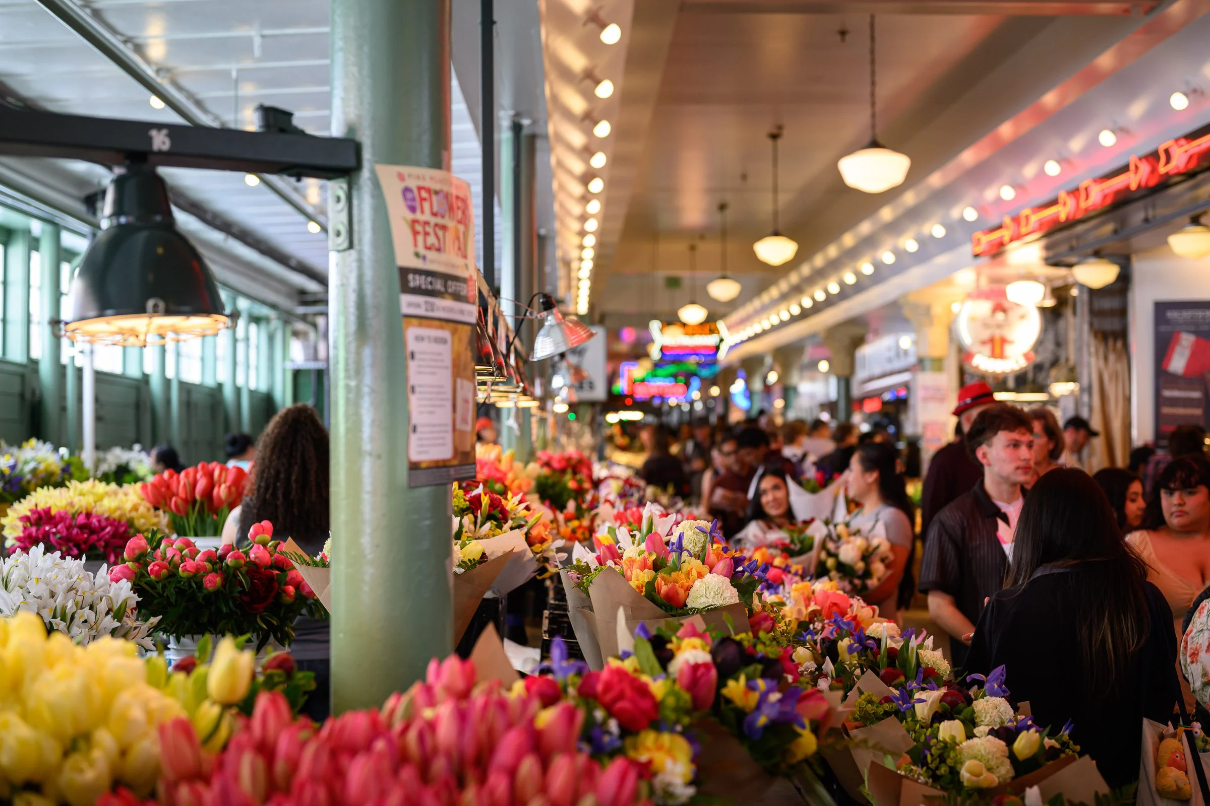 Indoor flower market filled with colorful bouquets of tulips, roses, and other flowers, with a crowd of people shopping amid bright overhead lighting and neon signs.