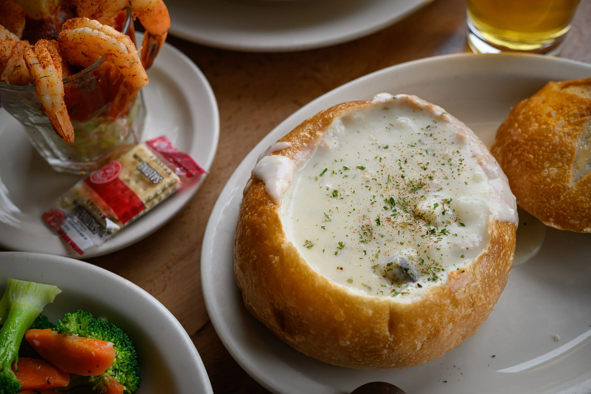 A bread bowl filled with creamy clam chowder sprinkled with herbs, alongside a plate of fried shrimp in a glass, and a small packet of crackers. A bowl of broccoli and carrots is partially visible.