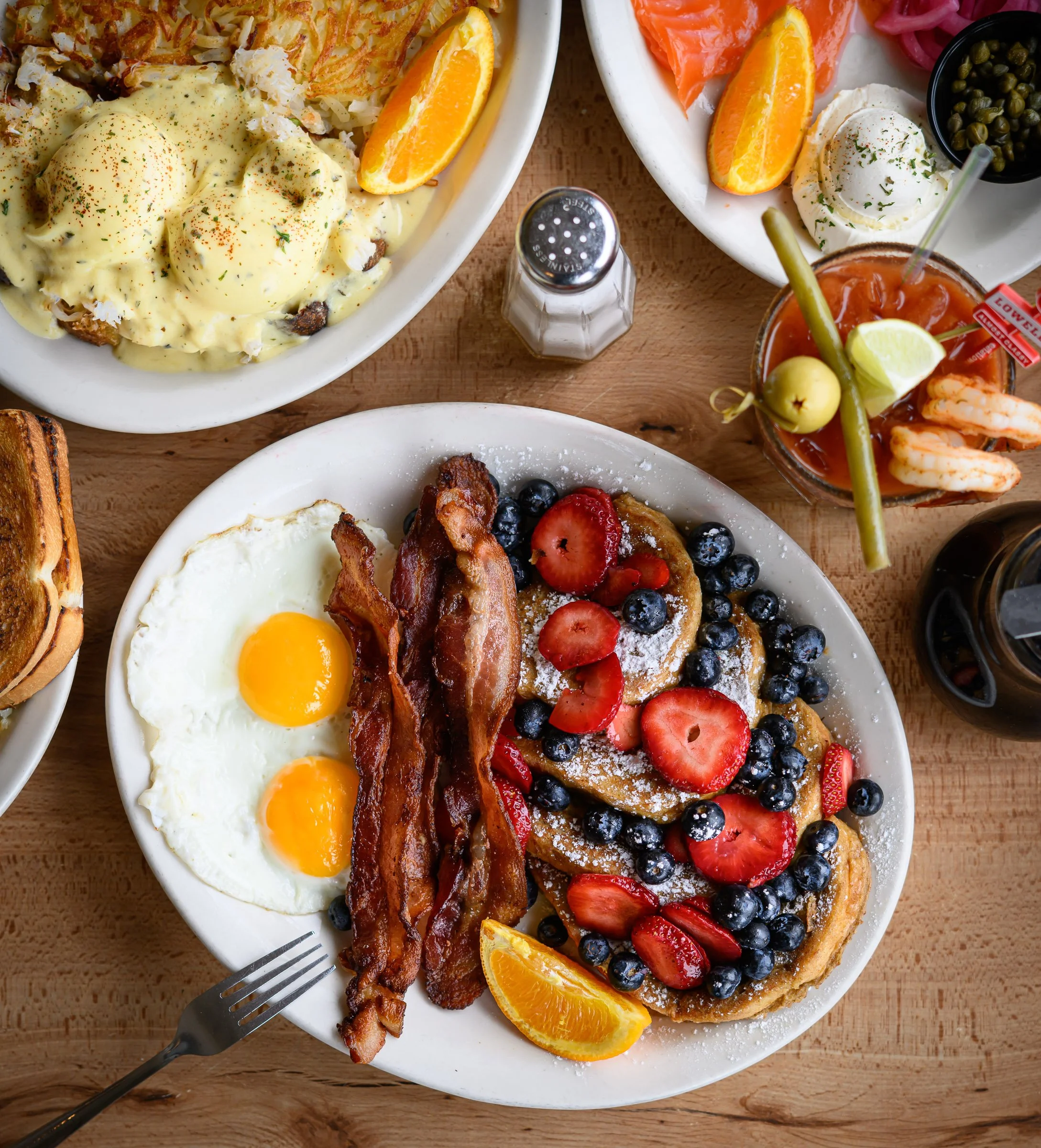 A breakfast plate with sunny-side-up eggs, bacon, and a waffle topped with strawberries and blueberries. An orange wedge is also on the plate. There is a fork on the plate's edge.