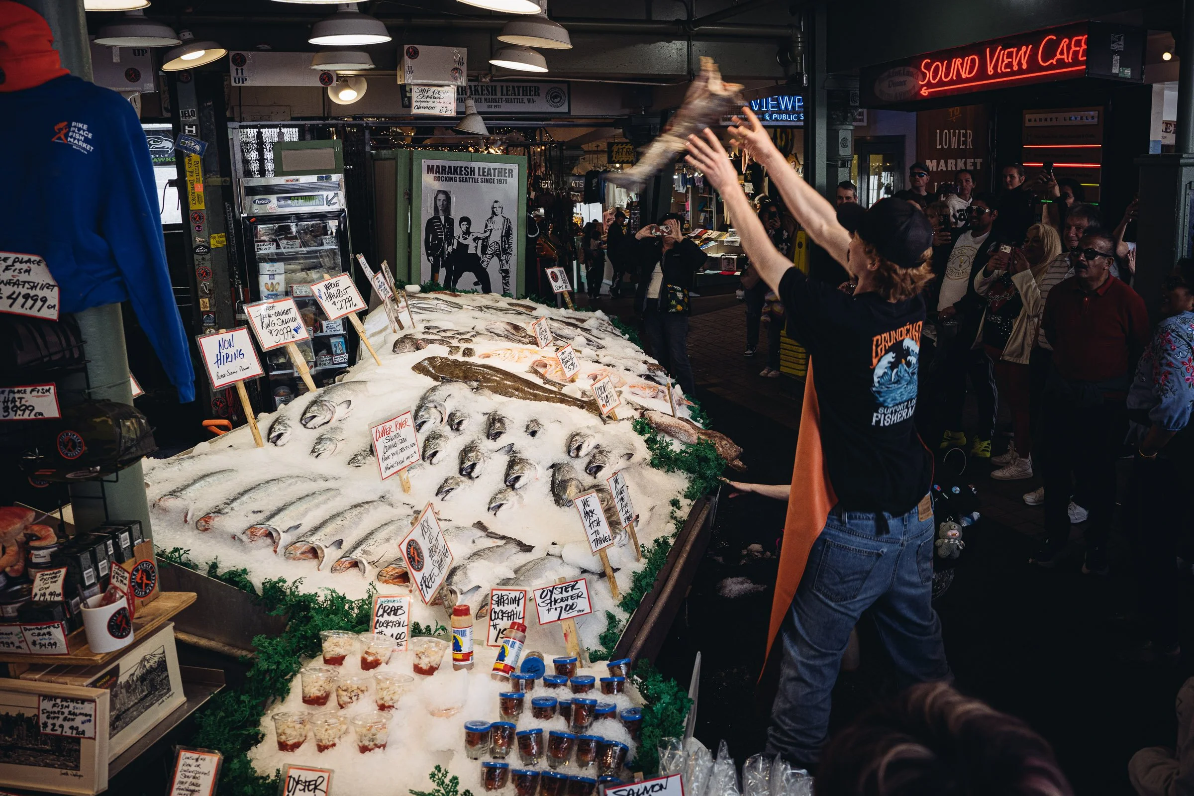 A fish market stall with various fish and seafood on ice, including salmon, crab, shrimp, and oysters, with price tags and signs. People are shopping and a person in the foreground is tossing fish, with a crowd of onlookers in the background.
