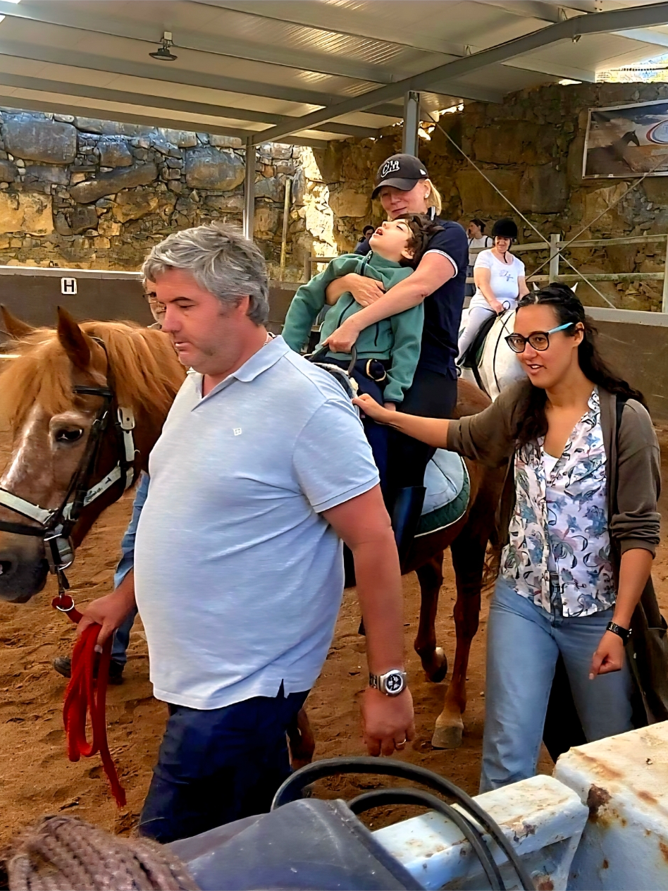 People riding horses in an indoor arena with stone walls and a roof, a young boy sitting on a horse with a woman holding him, and two adults walking nearby.