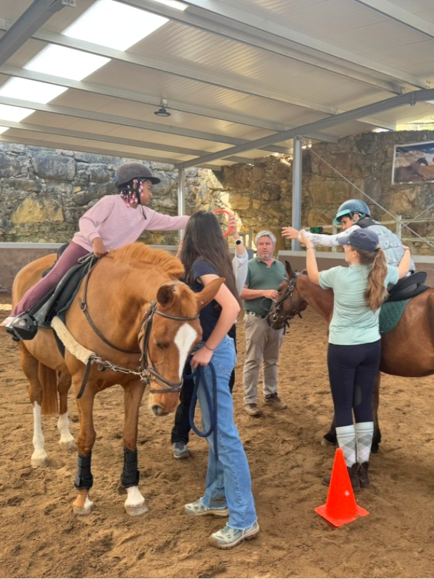 A girl in a pink shirt and helmet is sitting on a brown horse, giving a high five to another girl in a teal shirt and helmet who is on a different horse. Two women and a man are standing nearby, with the woman holding the reins of the first horse. The scene takes place in an indoor riding arena with a sandy floor and rocky walls, and a bright skylight overhead.