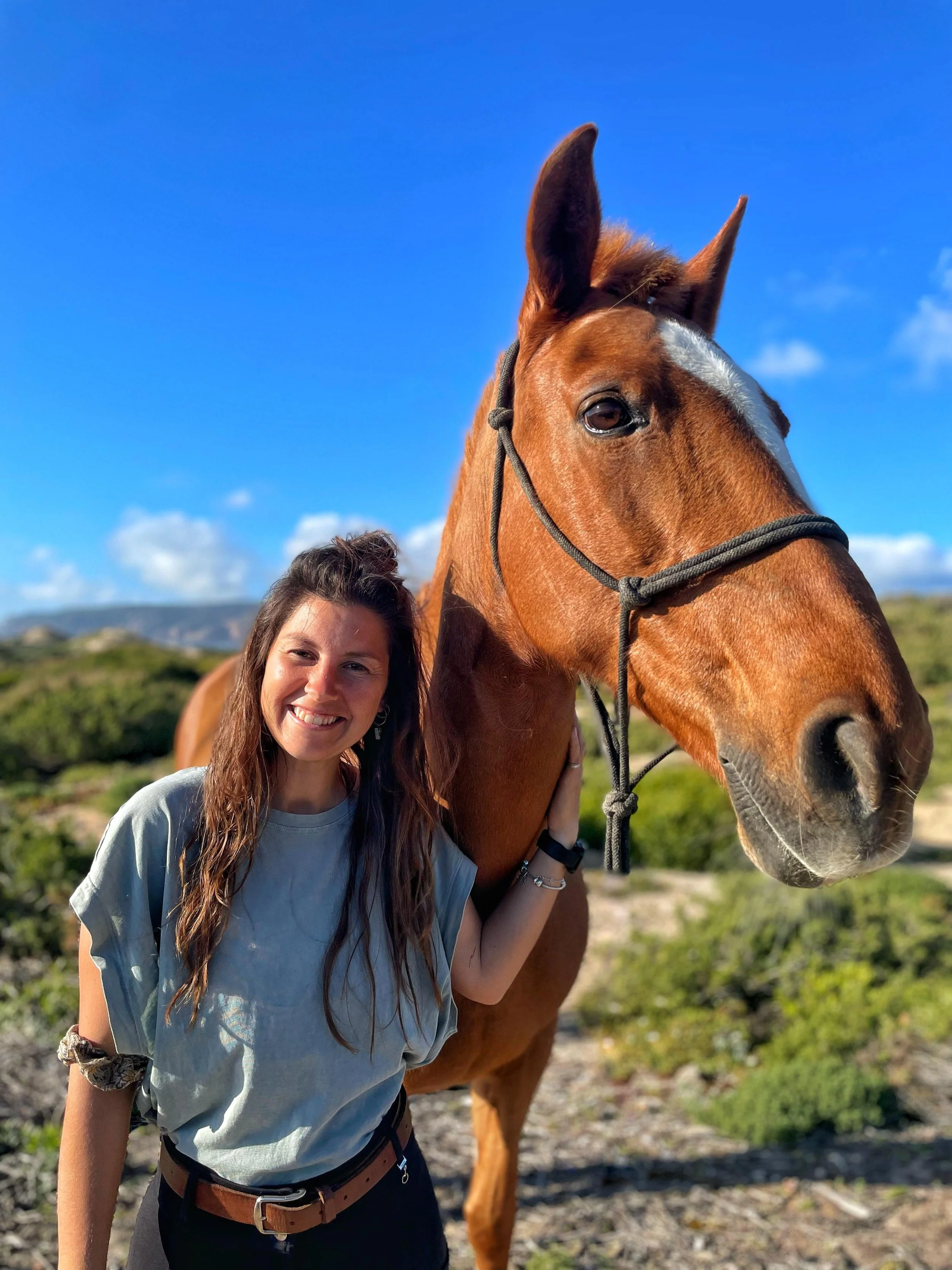 A woman with long brown hair smiling and standing close to a brown horse with a white stripe on its face, outdoors in a sunny field with blue sky and clouds.