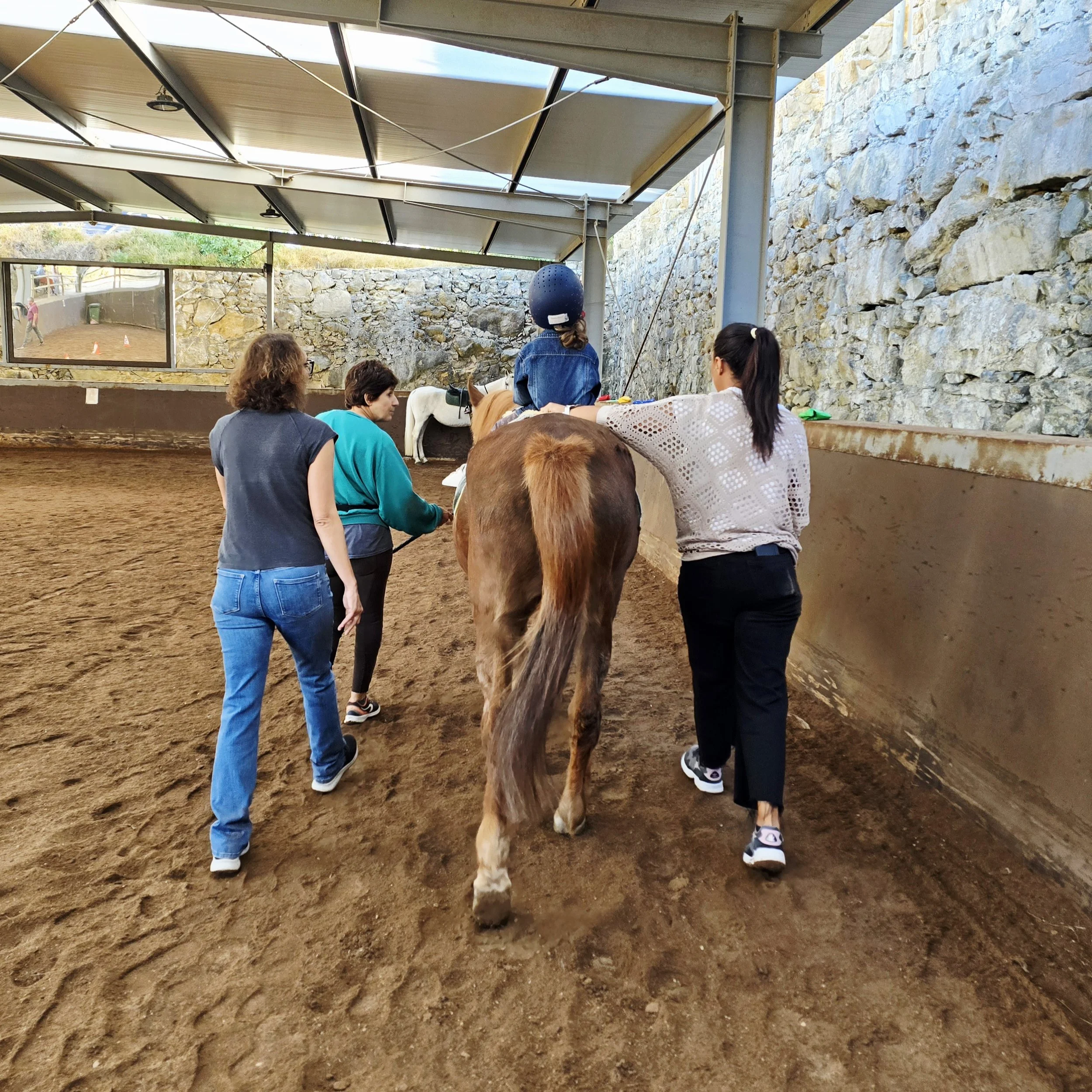 People and horses at an indoor riding arena with a stone wall and a large mirror.