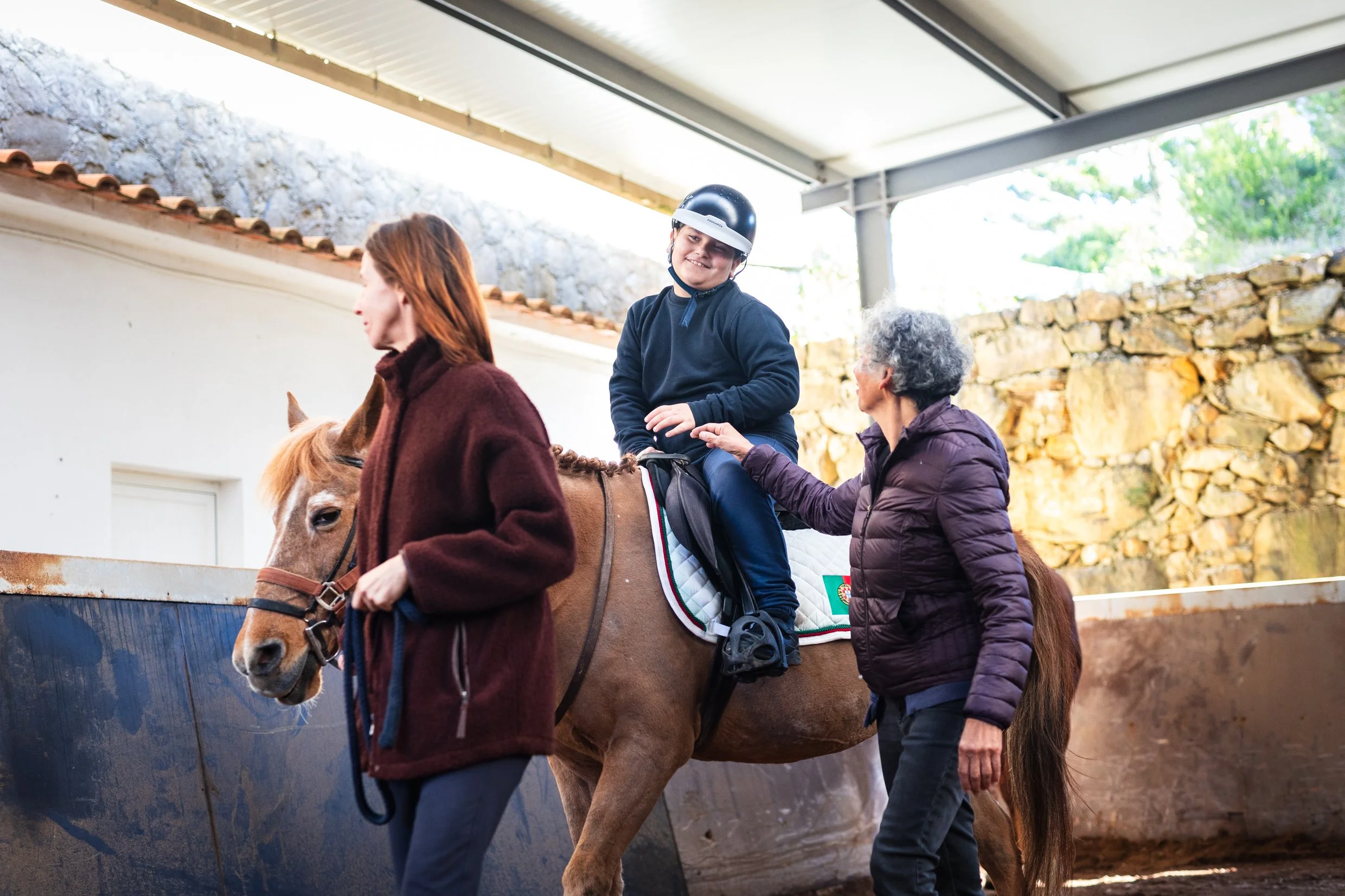 A child wearing a helmet riding a brown horse with an adult guiding it, in a covered outdoor area with stone wall and trees in the background.