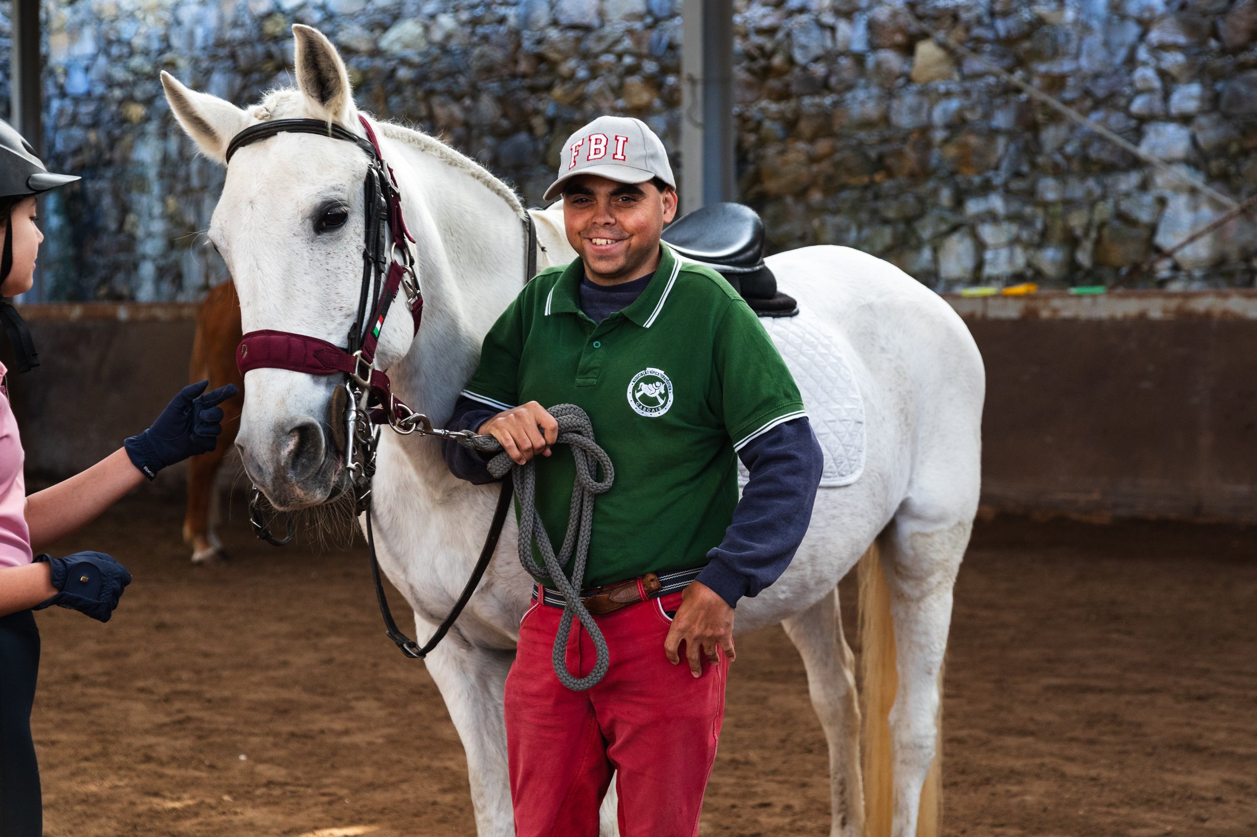 A man wearing a gray FBI cap and a green polo shirt with a horse riding emblem, smiling while holding the reins of a white horse with a bridle. A child with a helmet touches the horse's face. The background is an indoor riding arena with a stone wall.