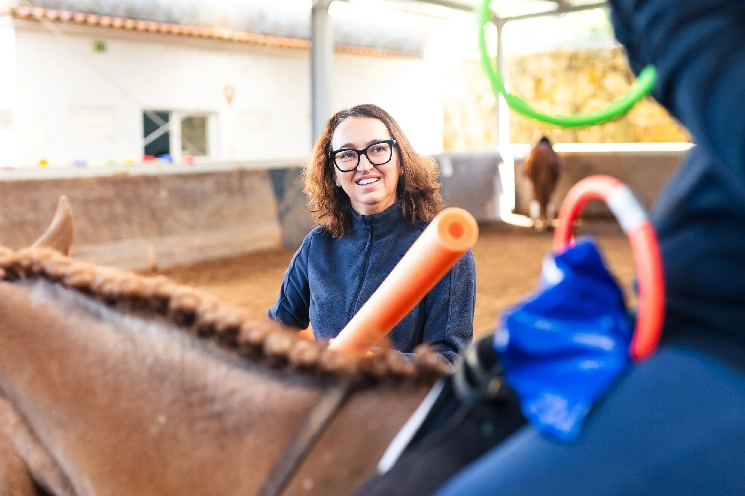 Smiling woman with glasses talking to person in a blue jacket at an outdoor petting zoo or farm exhibit, with animal enclosures and a barn in the background.