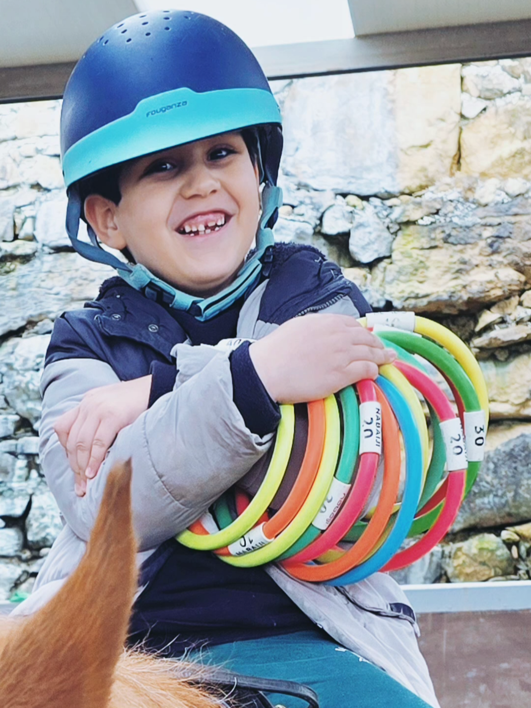 A young boy smiling with missing teeth, wearing a helmet, holding colorful hoops, sitting outdoors near a stone wall.