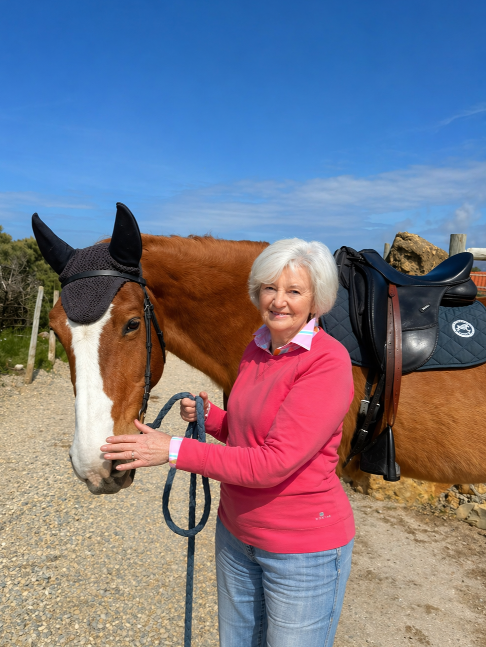 A woman with white hair holding a horse's reins and smiling, standing outdoors on a clear day with blue skies. The horse is brown with a white face and is wearing a black ear bonnet and saddle.