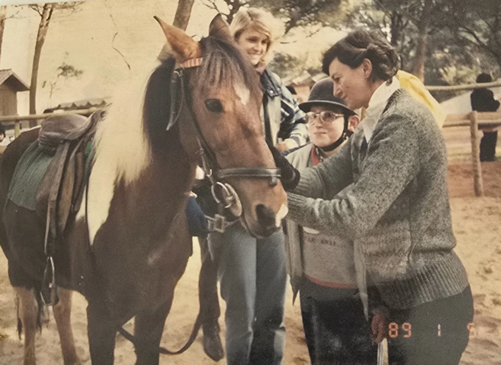 A man and a girl are petting a horse at an outdoor farm. The girl is smiling and has blonde hair, the man has dark hair and is wearing a gray sweater. Other children and a fence are visible in the background.