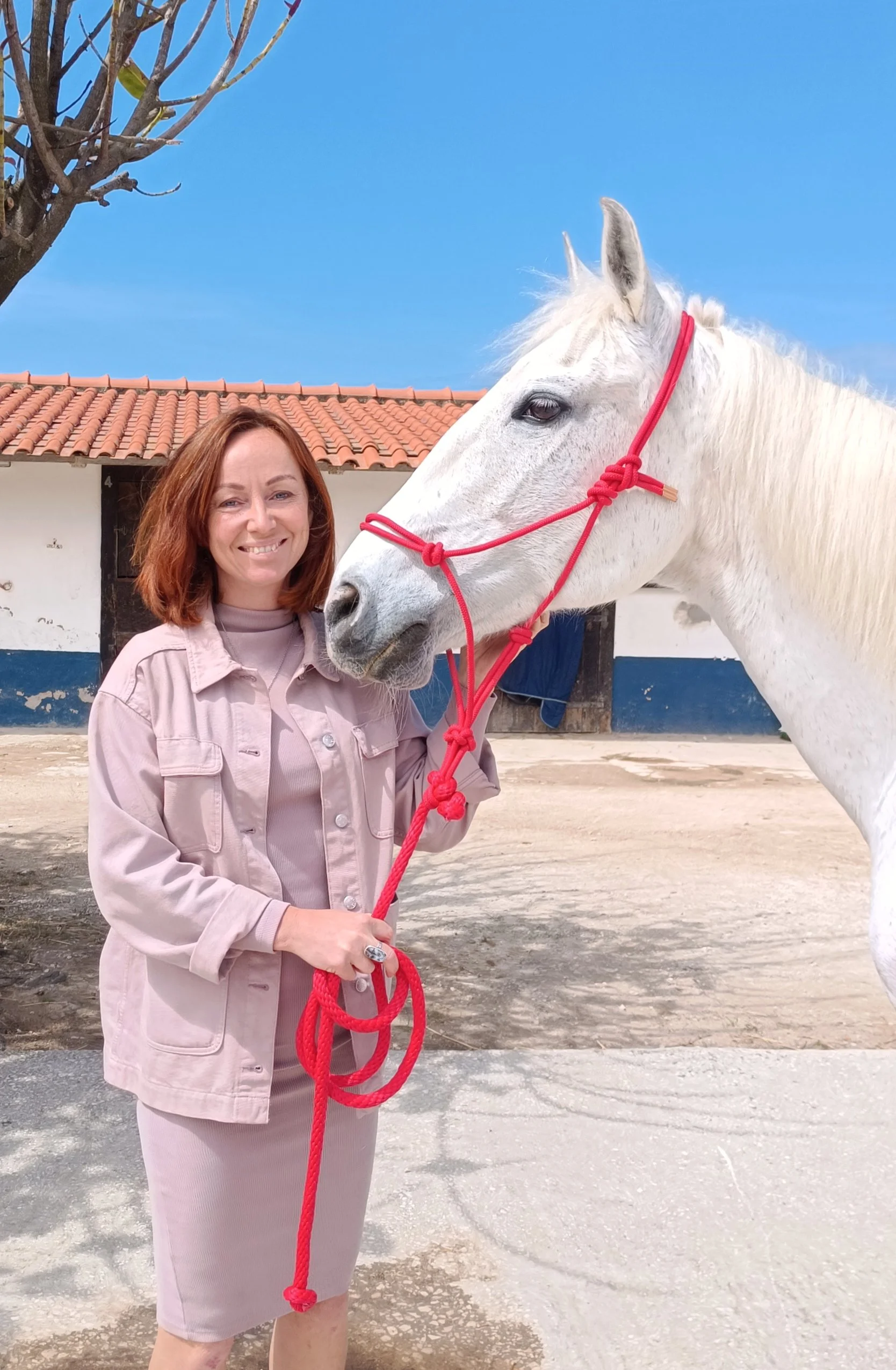 A woman with shoulder-length red hair wearing a pink jacket and a matching skirt, smiling and holding a red rope. She stands beside a white horse with a red halter, outdoors in front of a building with a terracotta tiled roof and a blue sky.