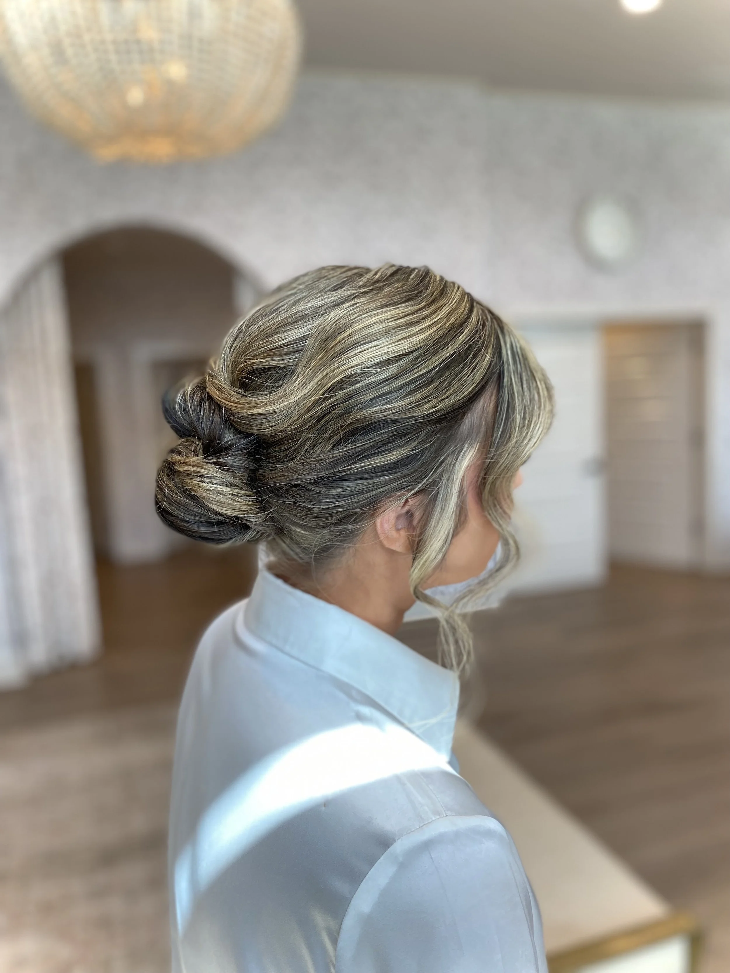 A woman with blonde hair styled in an elegant updo, braided and gathered at the nape of her neck, wearing a white garment in a well-lit room.