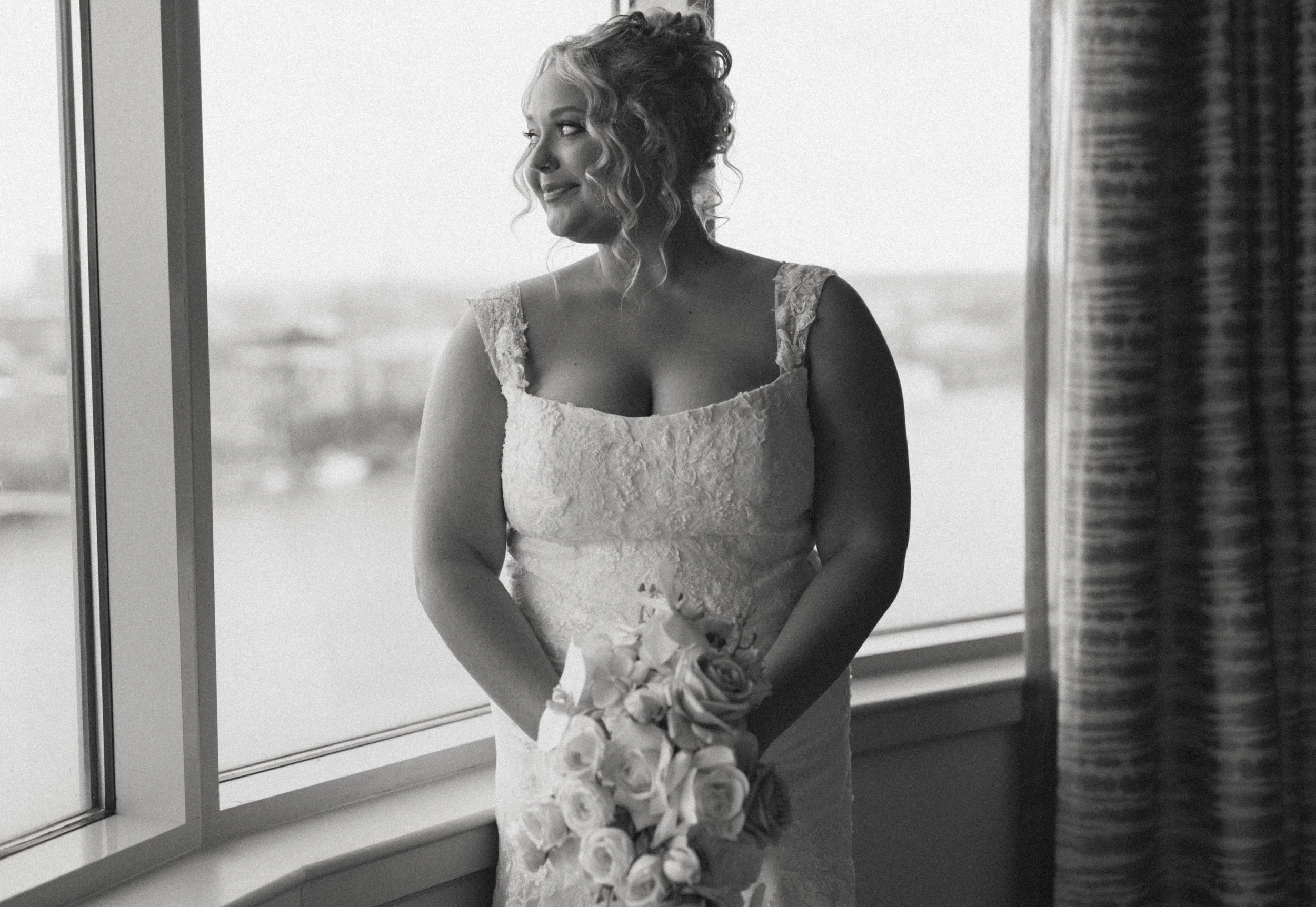 A woman in a lace wedding dress holding a bouquet of flowers, standing by a window and looking out with a smile.