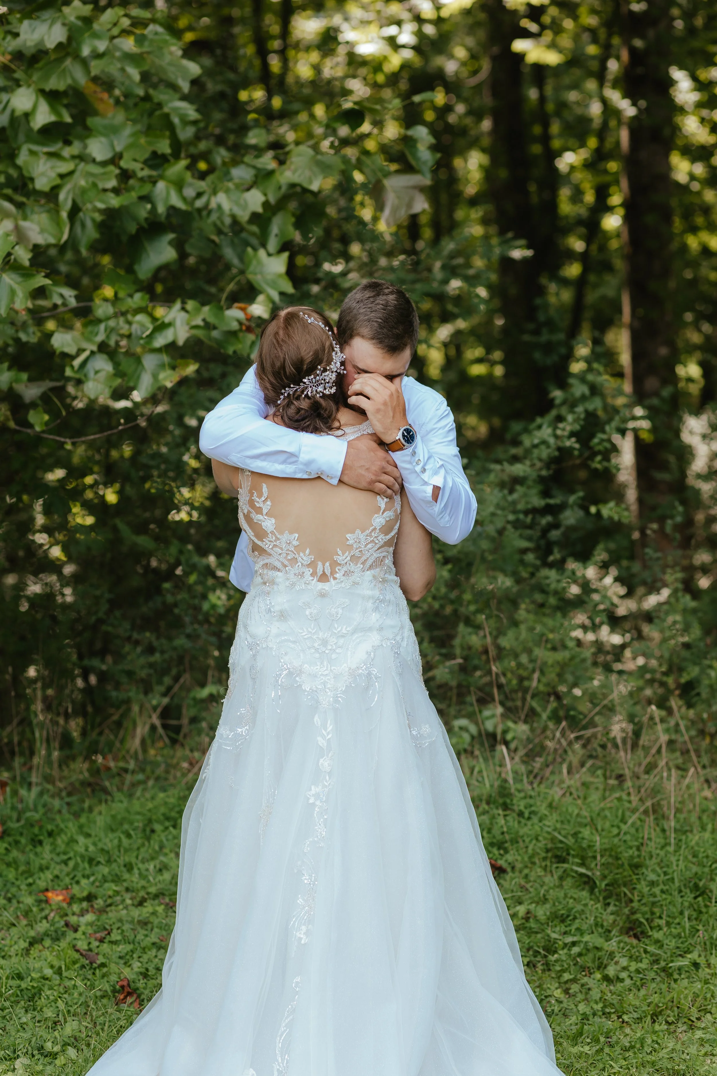 A bride and groom embrace outdoors surrounded by greenery, with the groom covering his face and the bride's back visible in a white wedding dress.