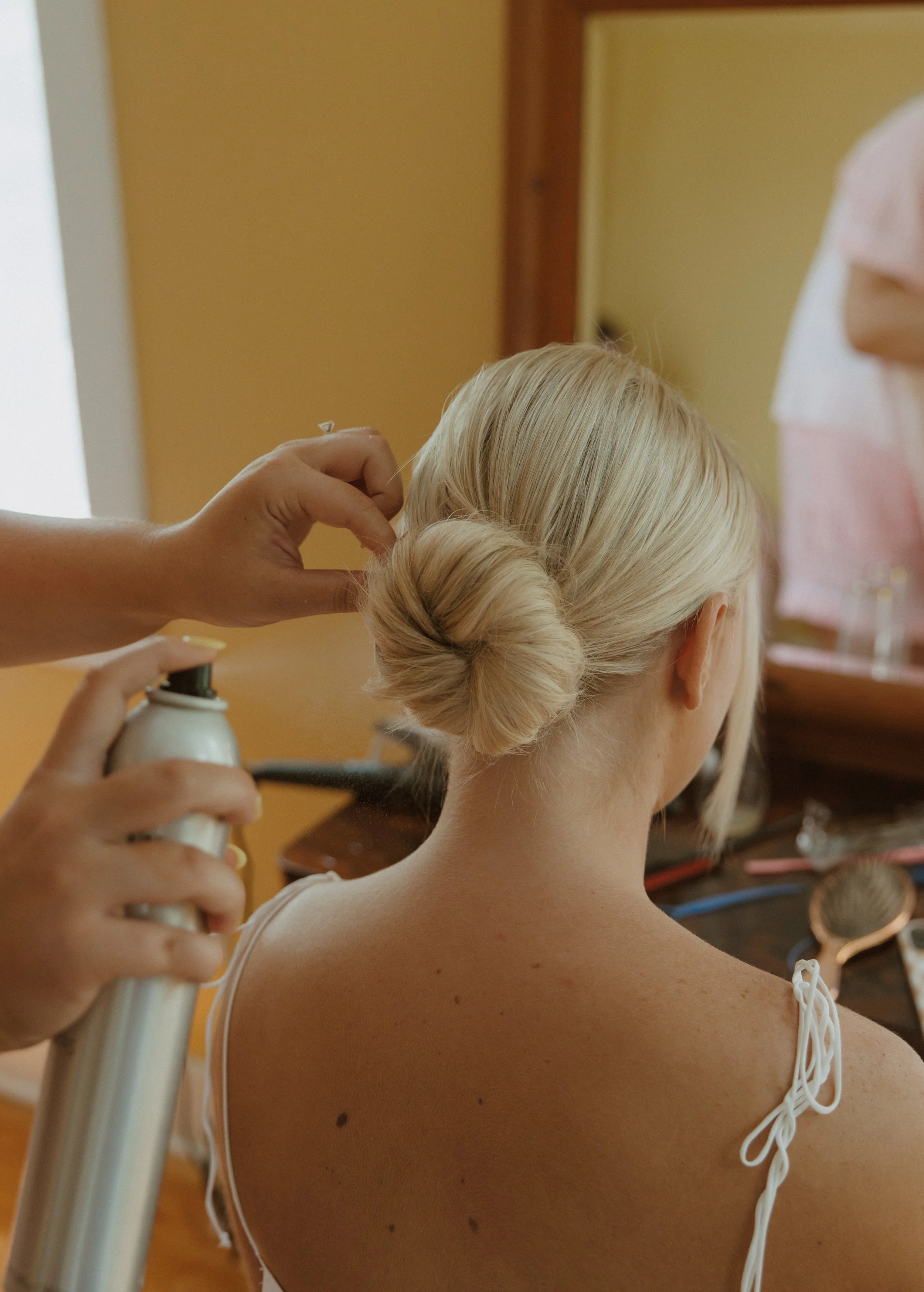 A woman with blonde hair styled in an elegant updo, dressed in a white dress, is having her hair styled by a person holding a spray bottle and a hairpin, in a room with warm yellow walls and wooden furniture.