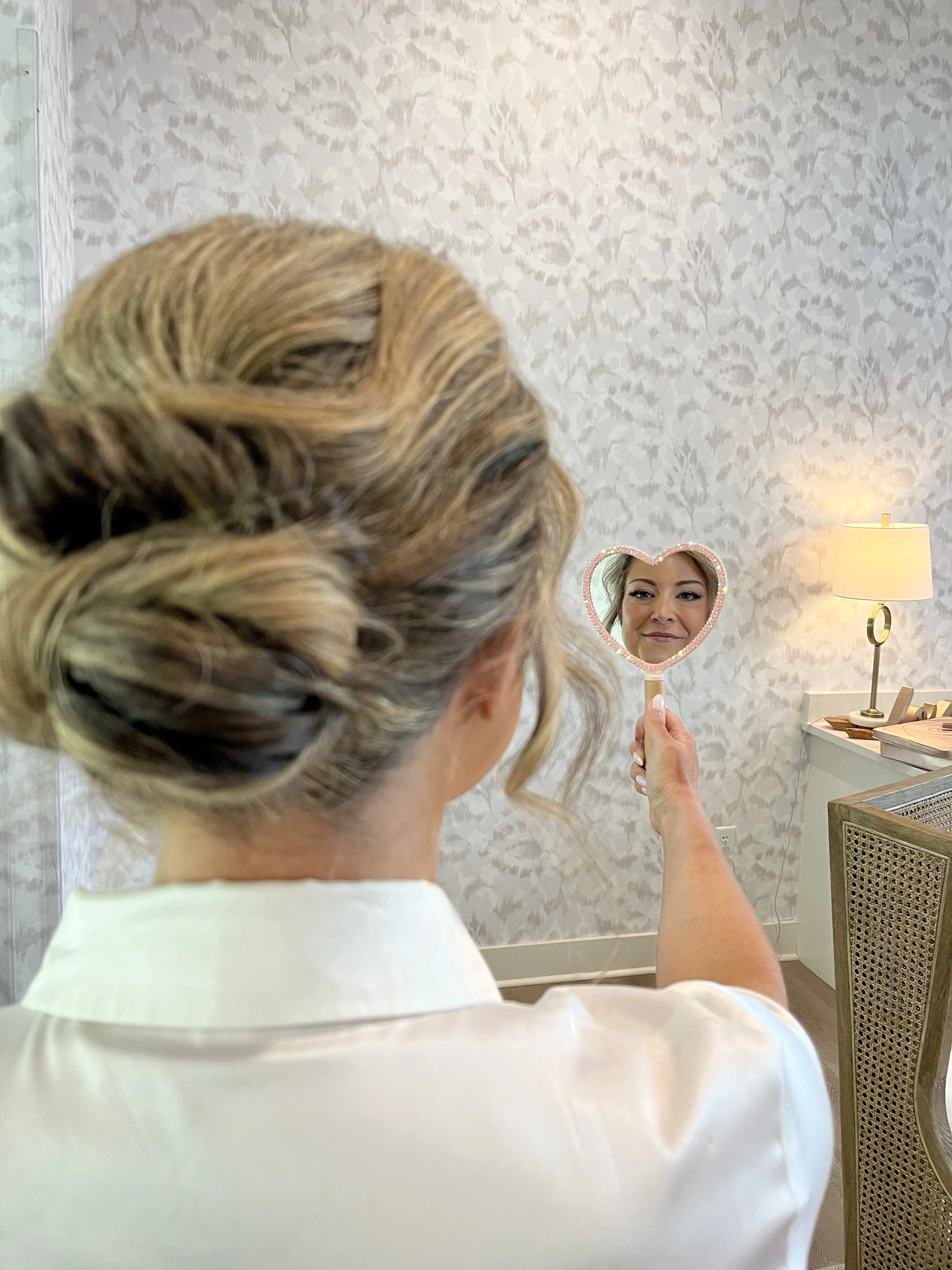 A woman with blonde, curled hair looking into a small, heart-shaped mirror held in her right hand, as she prepares or checks her appearance in a room with patterned wallpaper, a table lamp, and furniture.