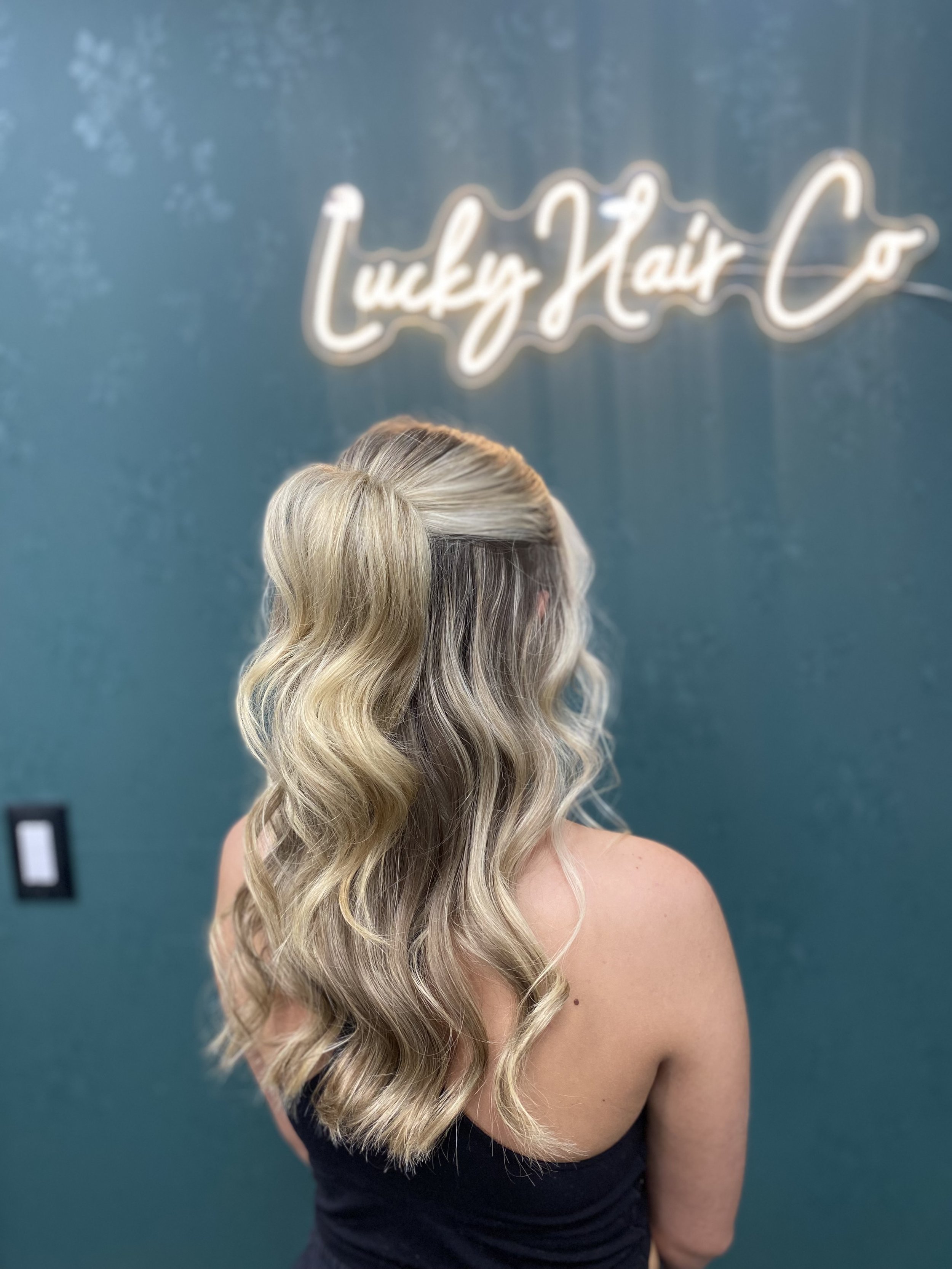 A woman with styled, wavy blonde hair in a loose ponytail, wearing a strapless black dress, standing against a teal-colored wall with a neon sign that reads 'Luxury Hair' in cursive.