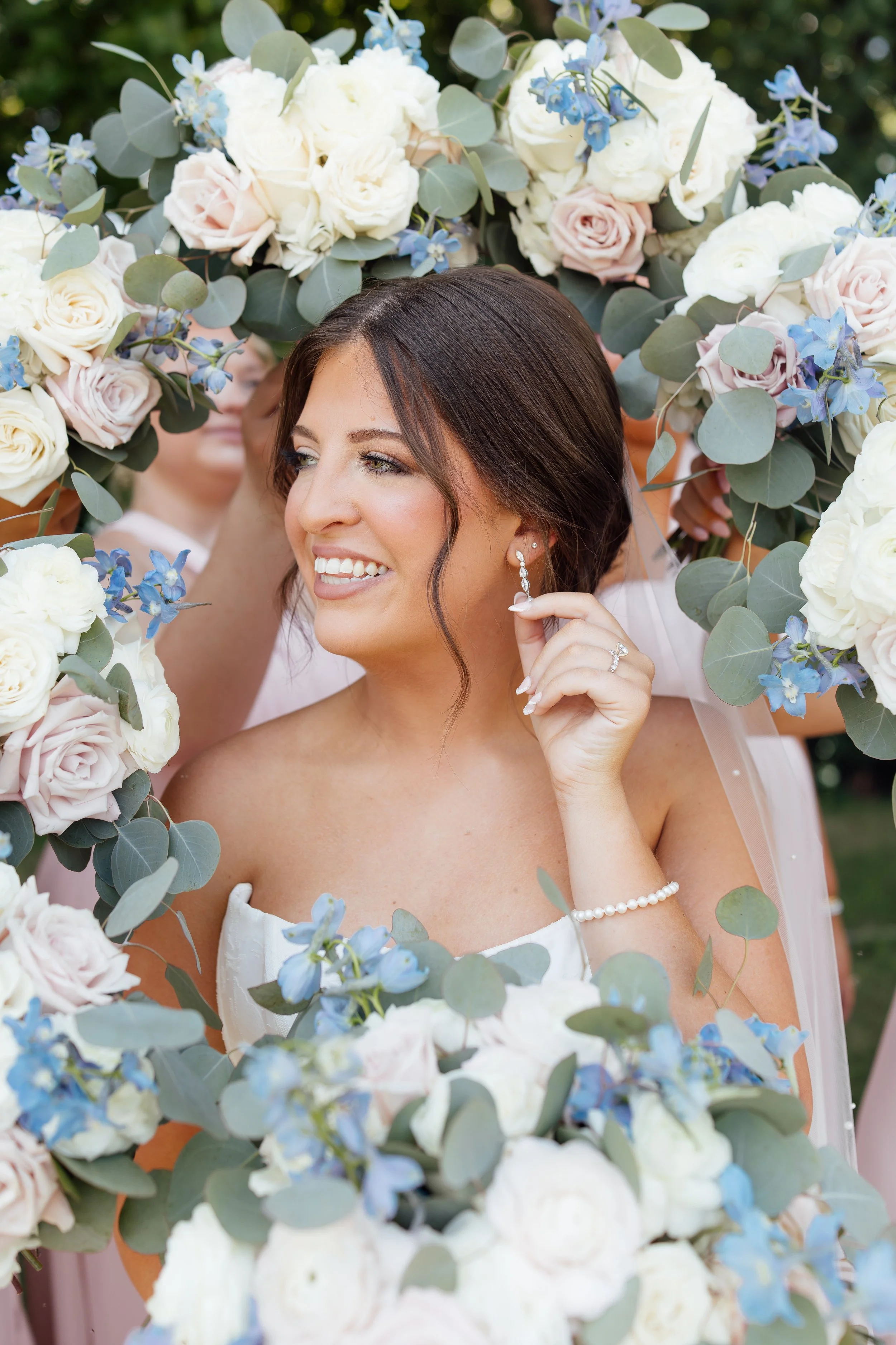 A bride with dark brown hair and earrings, smiling as she looks away, surrounded by a floral arch of white, blush pink, and blue flowers with green leaves, outdoors during the daytime.