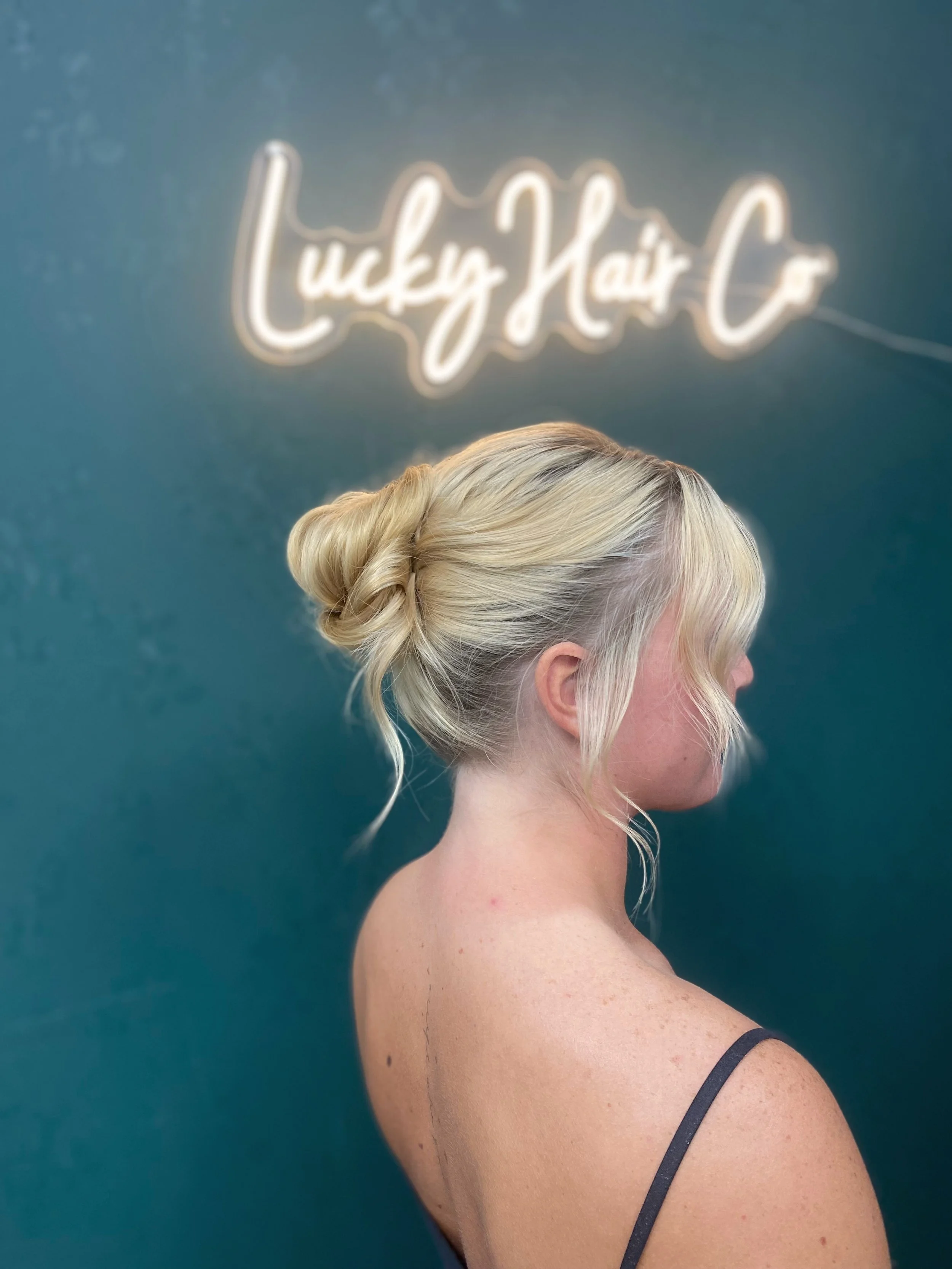 A woman with blonde hair styled in a loose bun, seen from the side, against a dark teal background with a neon sign that says 'Lucky Hair Co'.