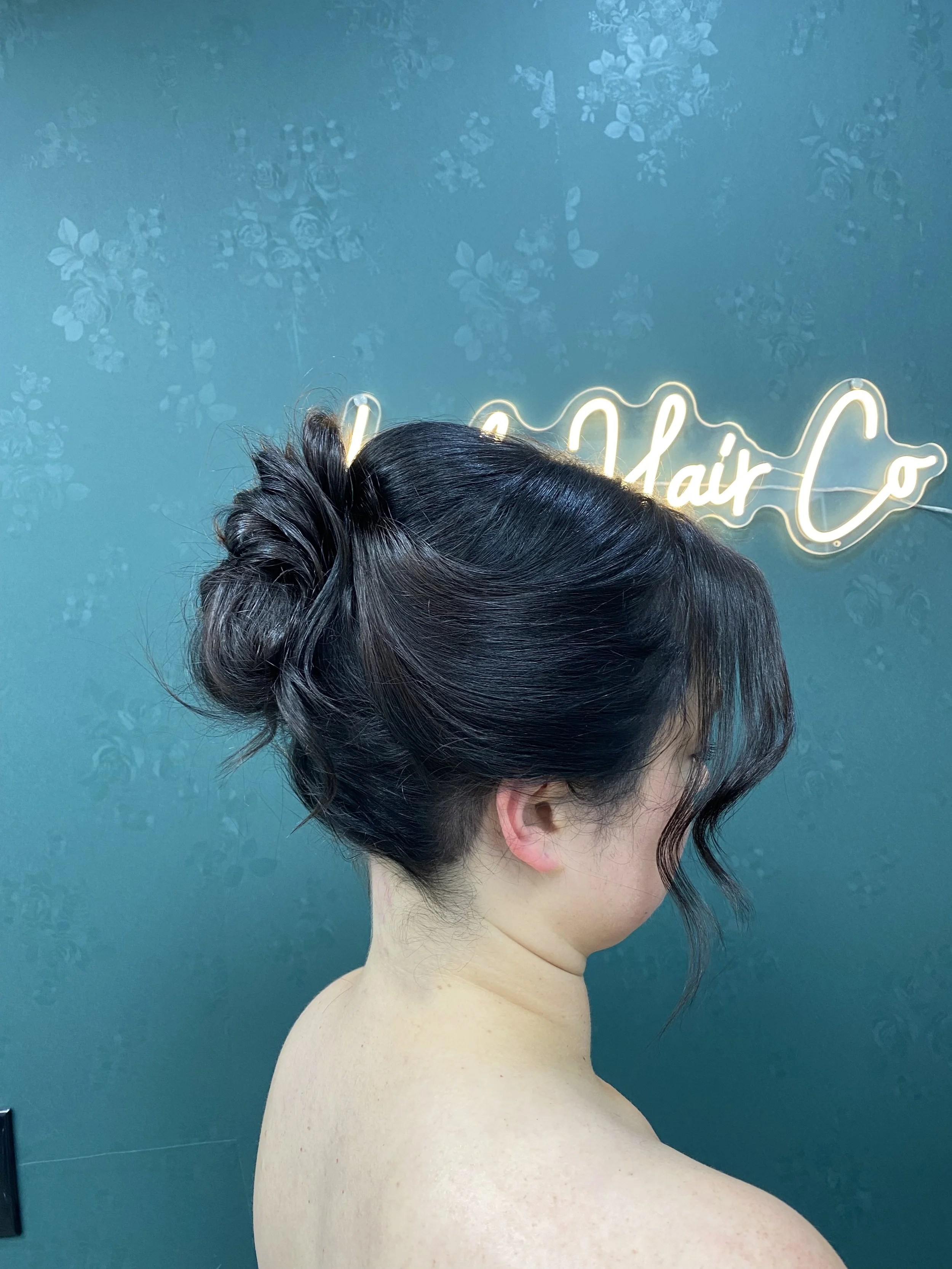 A woman with dark, styled hair in an elegant updo with loose curls framing her face, against a teal wall with floral patterns and a neon sign.