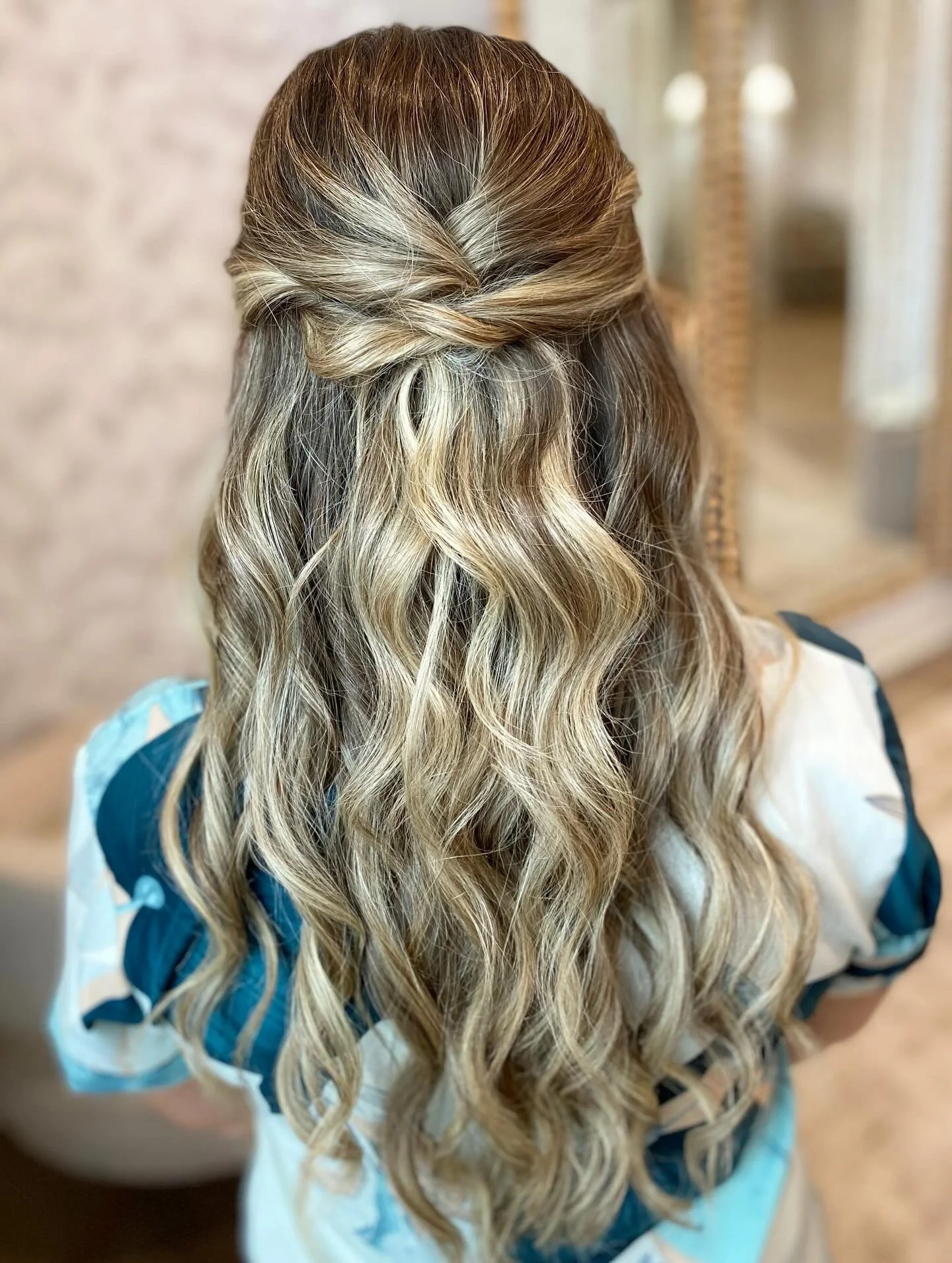 Back of a woman's head showing long, wavy blonde hair with darker roots styled with a half-up twisted hairstyle.