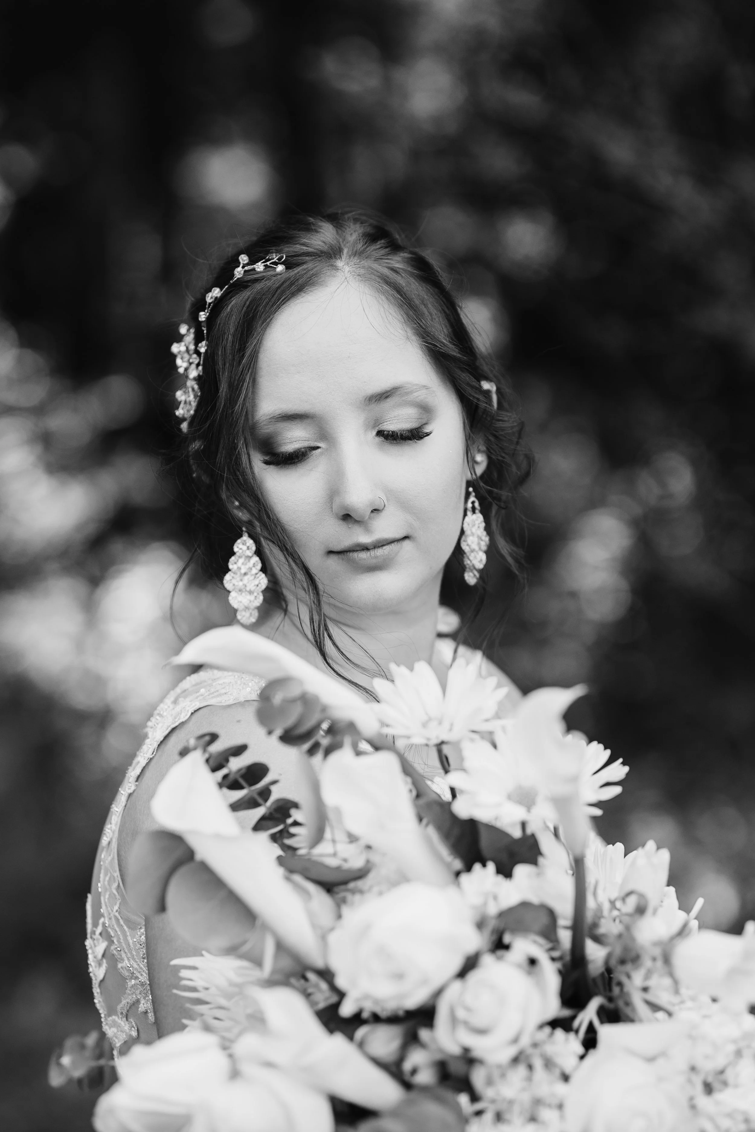A black-and-white photo of a woman with styled hair and ornate earrings, holding a bouquet of flowers, with a blurred natural background.