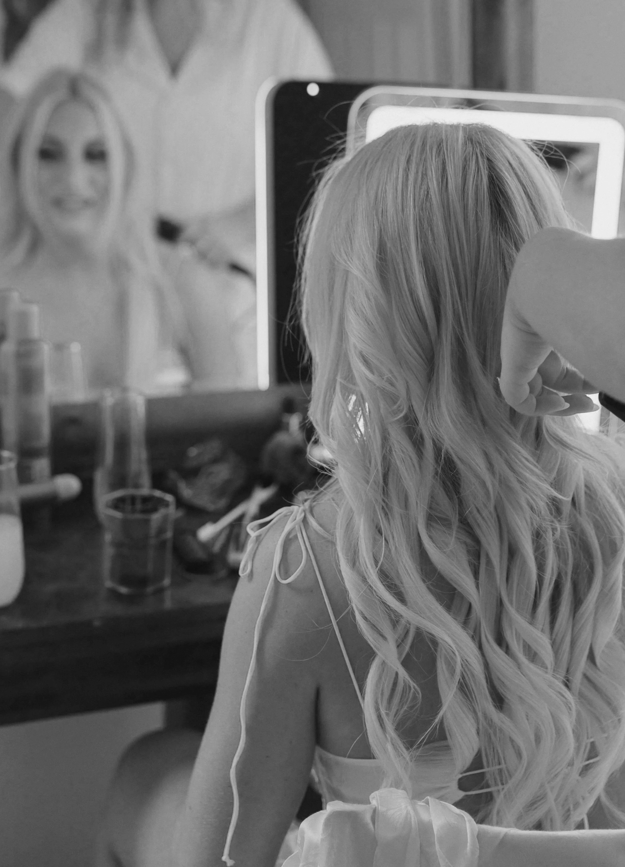 A woman with long, wavy blond hair sitting in front of a mirror, receiving a beauty treatment, with beauty products on the table in the foreground.