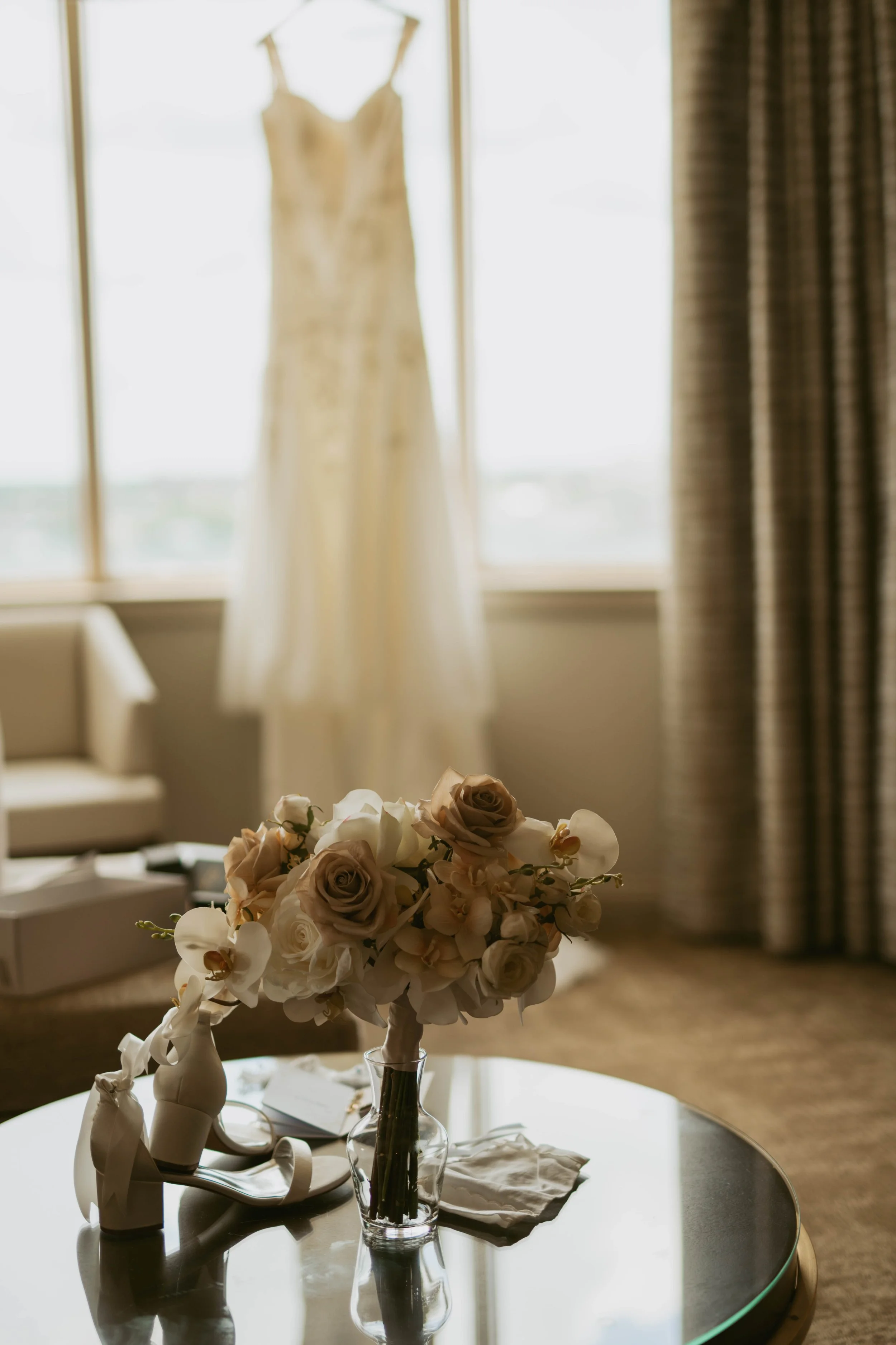A bouquet of white roses and orchids in a glass vase sitting on a table with a white cloth, with a blurred background of a window and curtains.