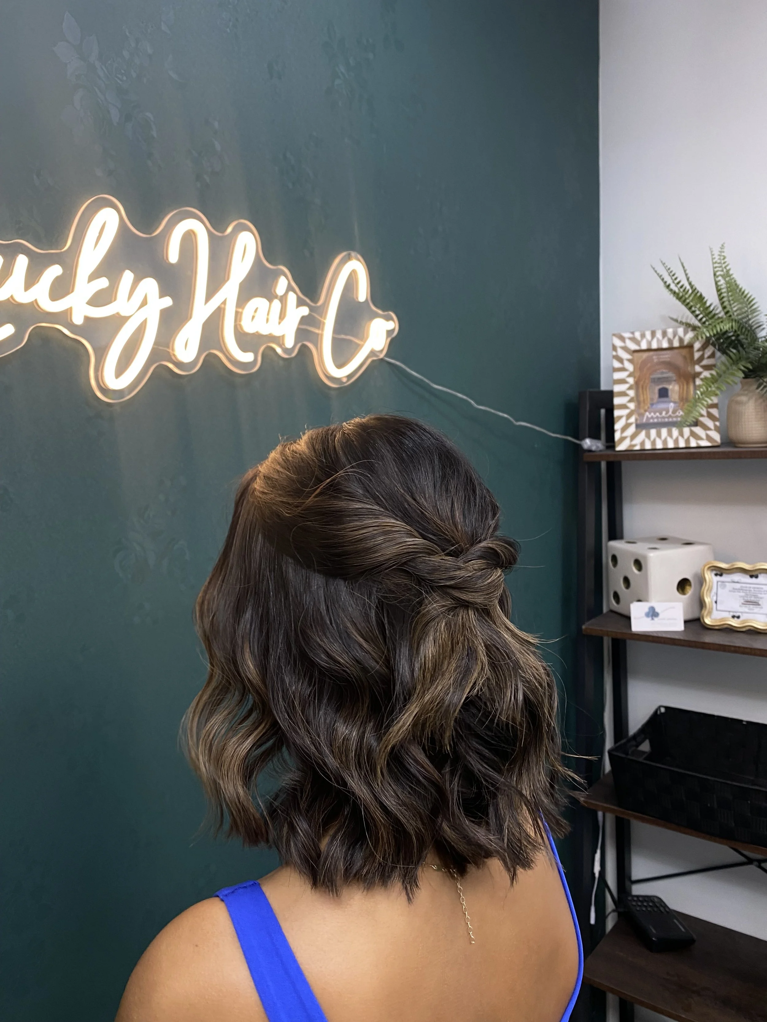 A woman with shoulder-length, wavy hair styled in loose curls, wearing a blue sleeveless top and a delicate gold necklace, standing in an indoor setting with a dark green wall and decorative items on shelves in the background.