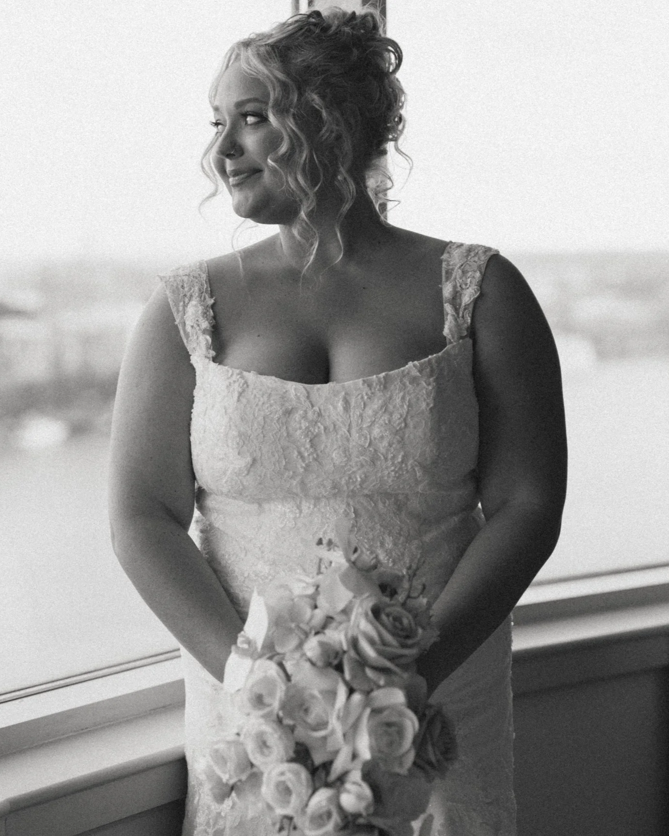 A woman in a wedding dress holding a bouquet of roses, standing near a window, smiling and looking to her left.